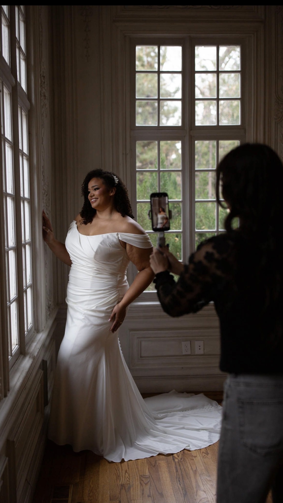 Woman in wedding dress poses by window, person filming with smartphone indoors, wooden floor and large window behind.