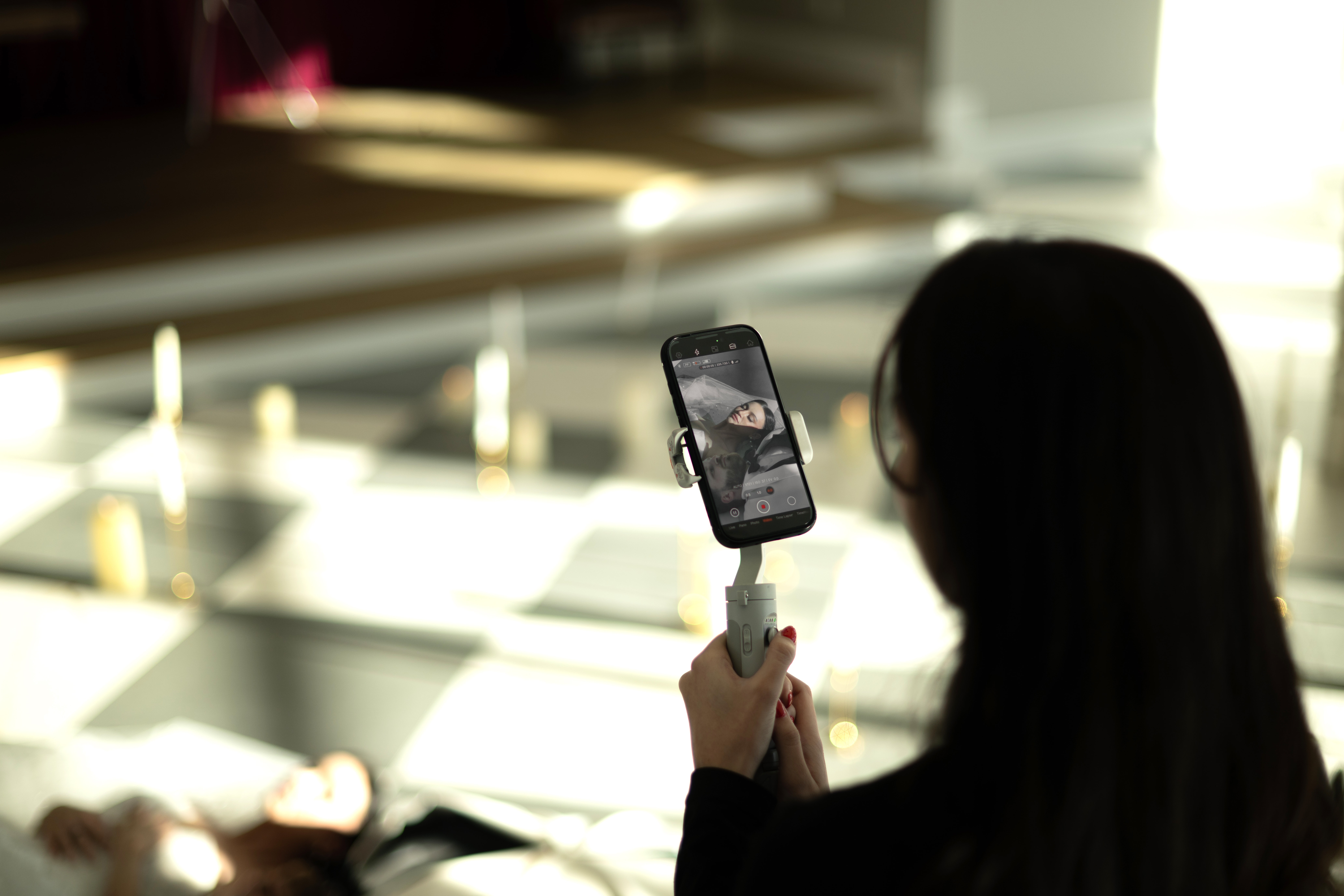 Person with long dark hair holding a smartphone, taking a photo or video indoors with bright light in background.