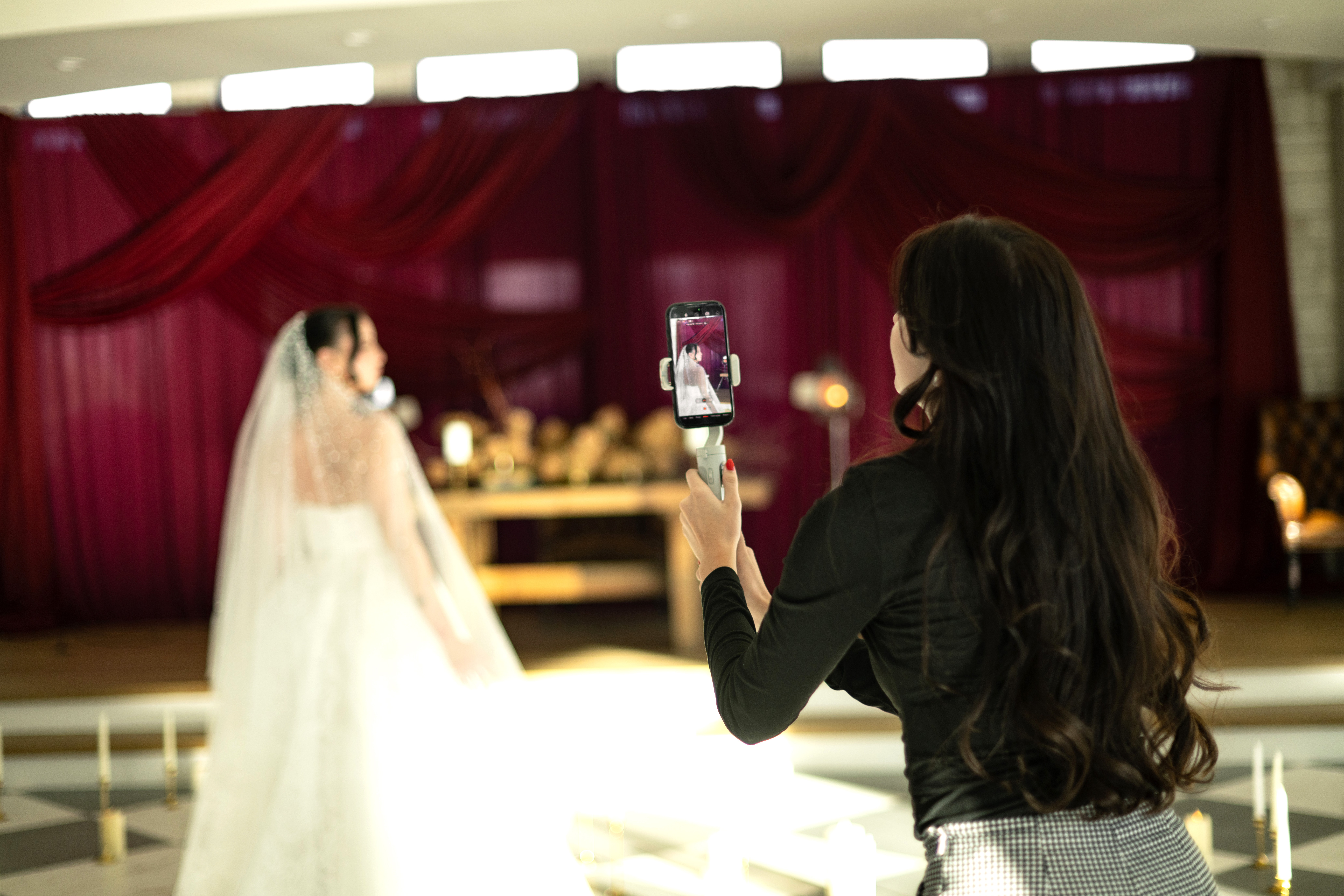 Person with long dark hair taking a photo of a bride in a wedding dress with a smartphone, indoors with red curtains.