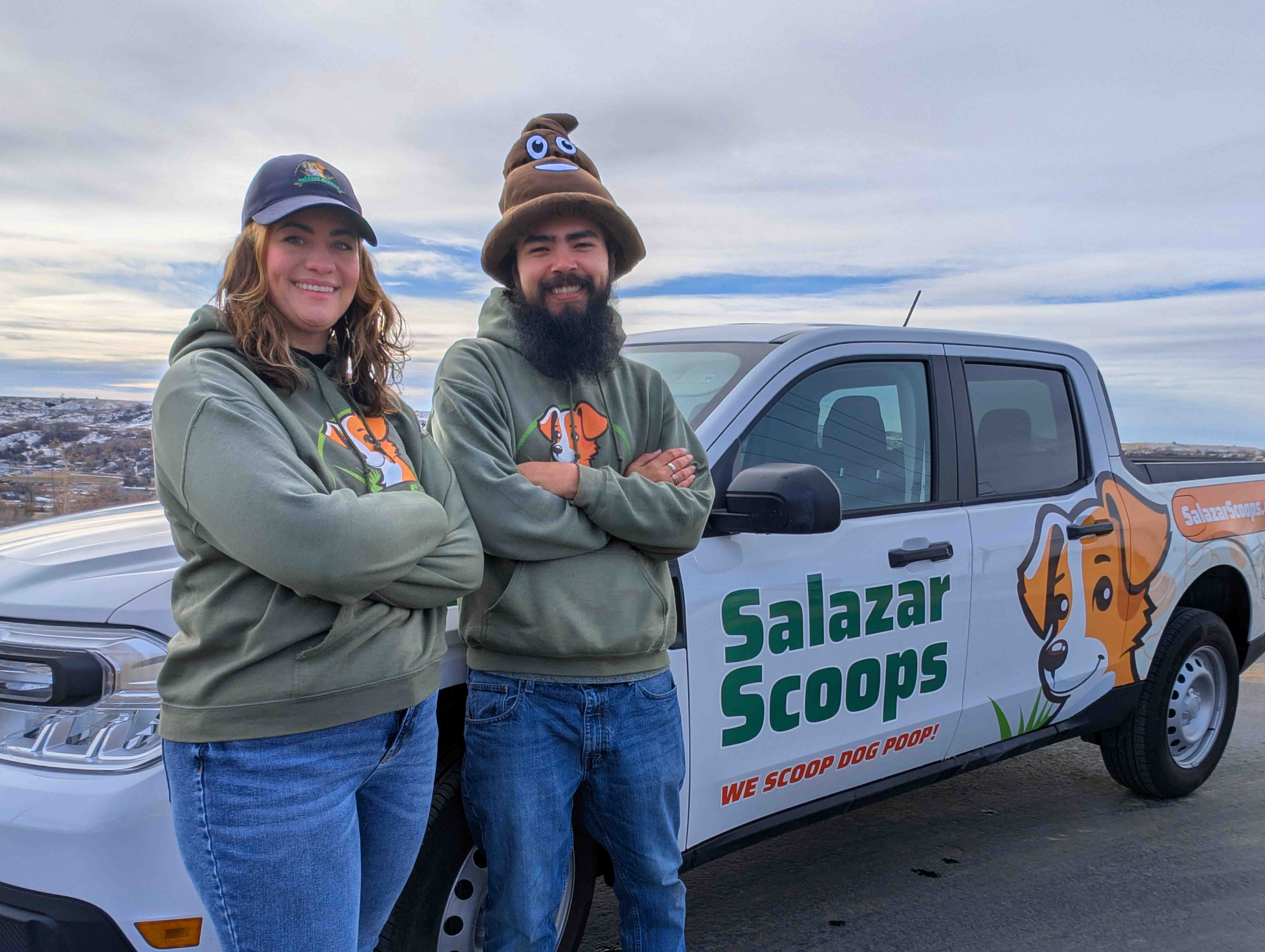 Two people stand in front of a pickup truck with a logo and text, outdoors on a cloudy day.