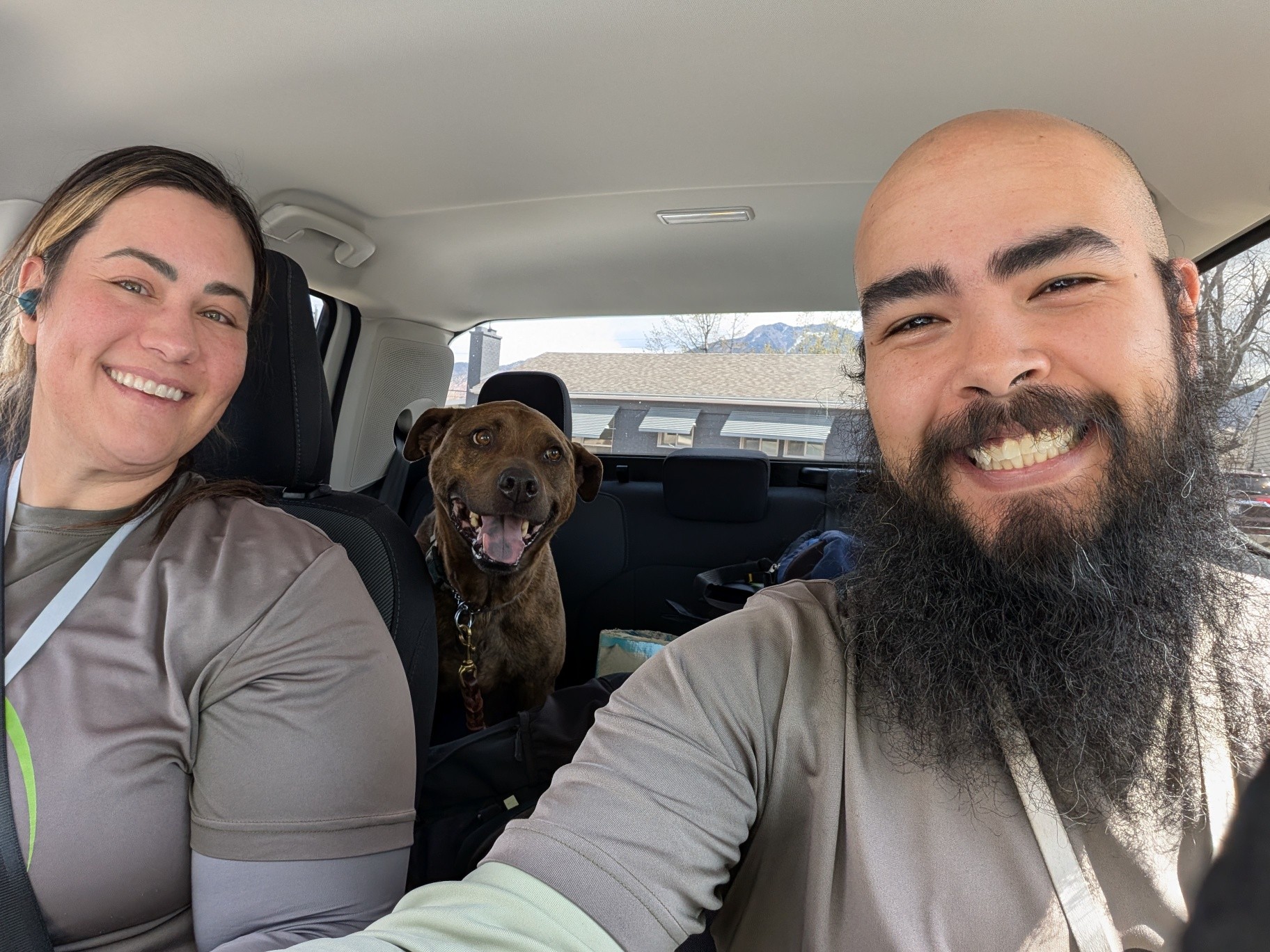 Two people and a dog sitting in a car, smiling at the camera, with a view of houses outside the window.