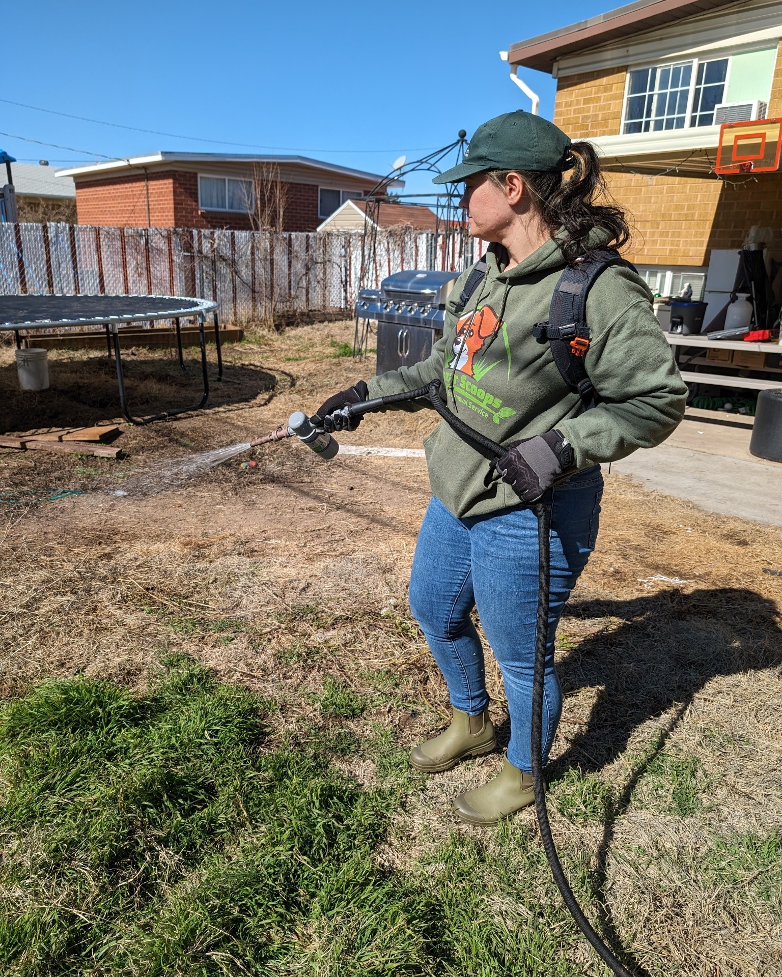 Person wearing a green cap, hoodie, and jeans watering grass in backyard, with trampoline and house in background.