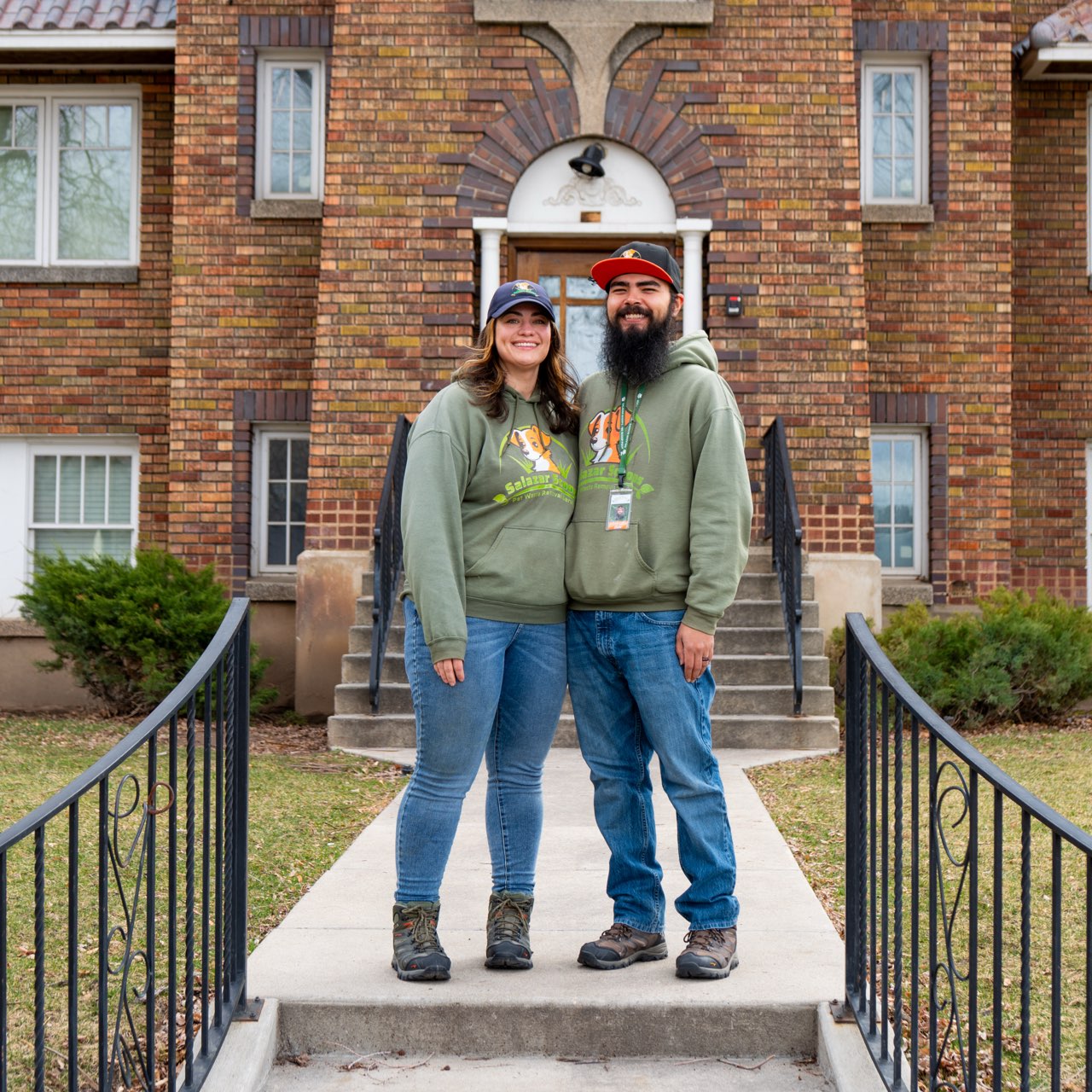 A smiling couple stands on a sidewalk in front of a brick building, wearing matching hoodies and jeans, with stairs behind them.