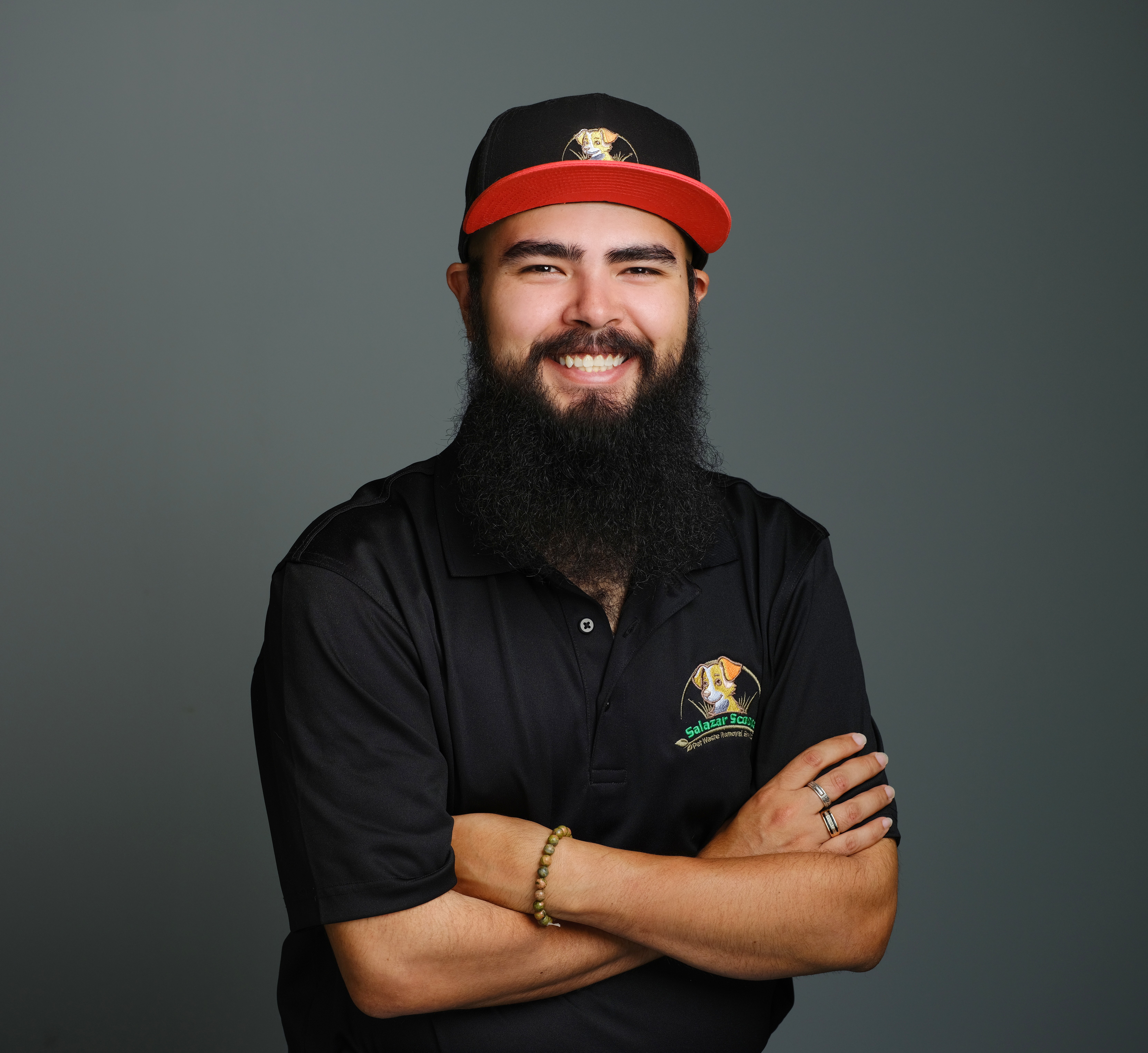 Smiling man with a beard wearing a black polo shirt and a black cap with a red brim, arms crossed.