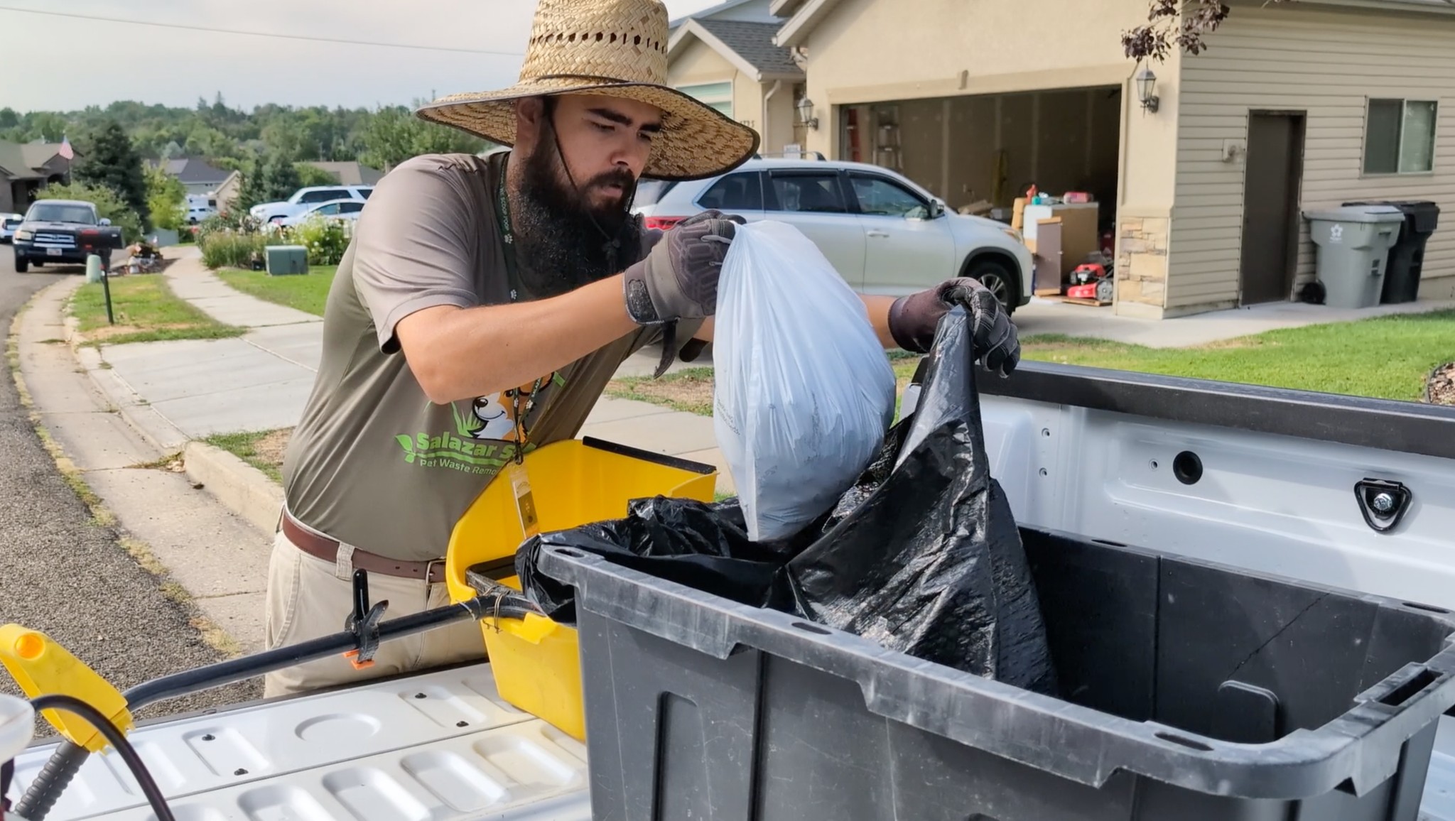 Man wearing a straw hat and gloves, disposing of trash into a large black bin outdoors.
