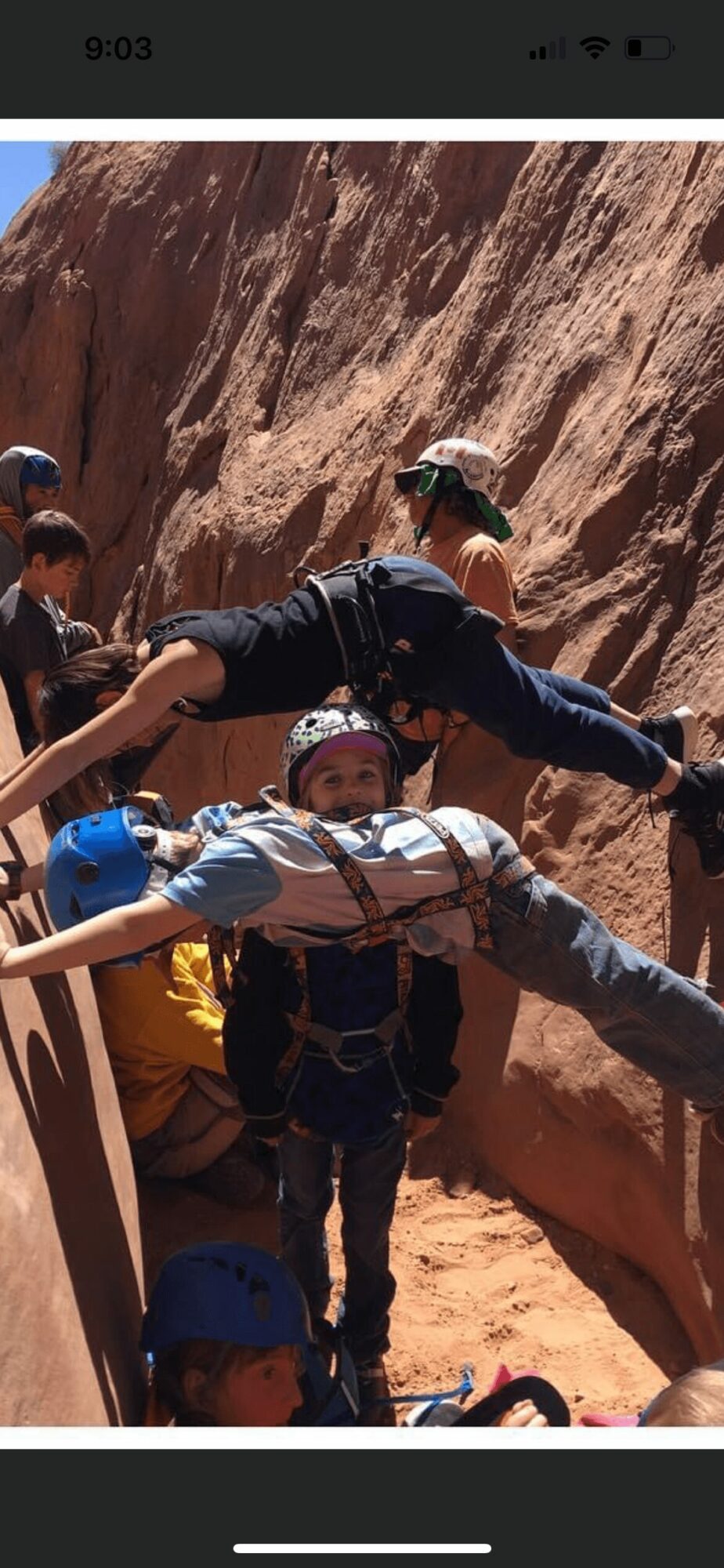 Group of people climbing a rocky outdoor area with a steep slope, some wearing helmets and harnesses.
