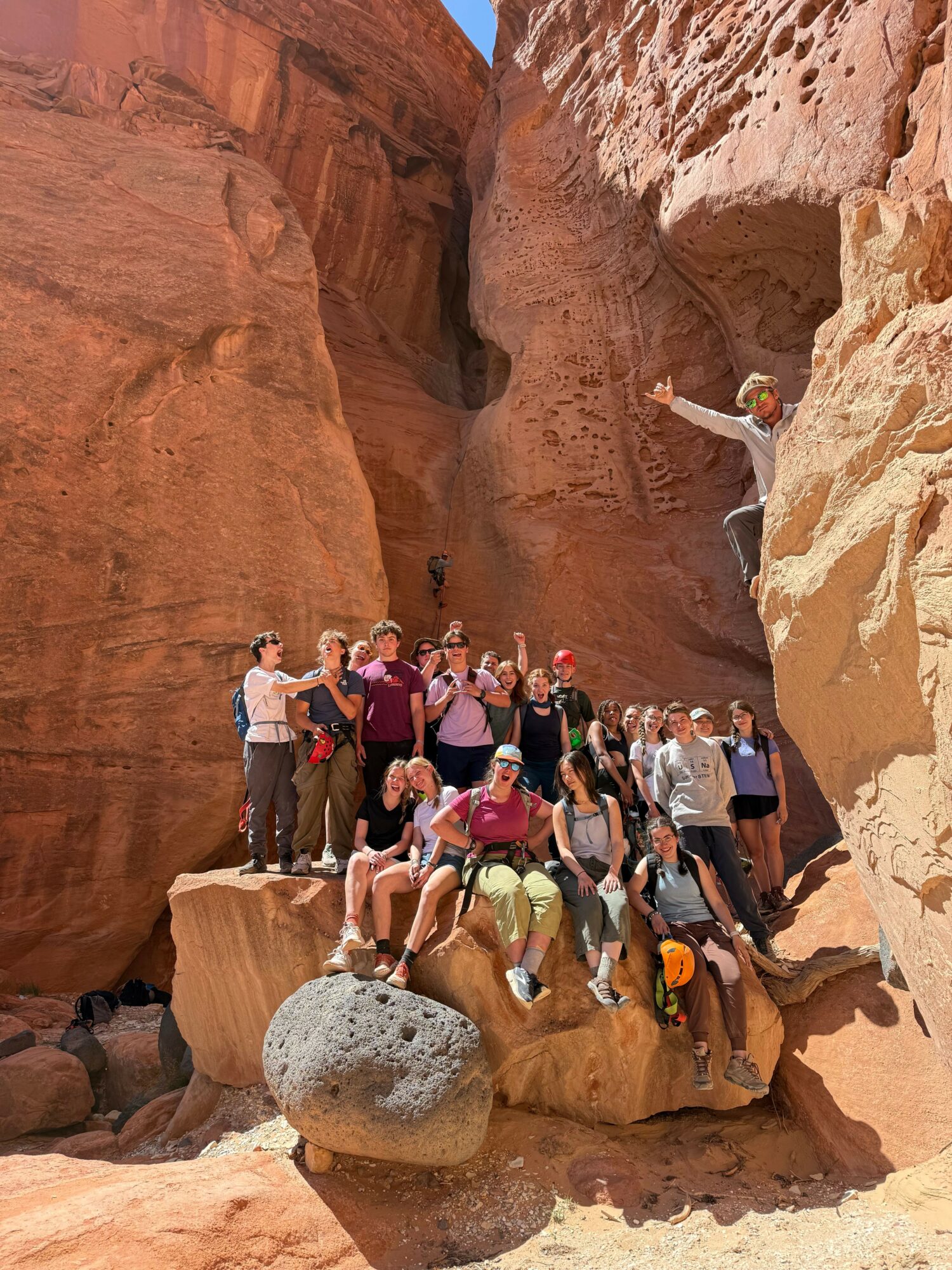Group of people inside a narrow canyon with reddish rock walls, some climbing and others standing, with a large boulder nearby.