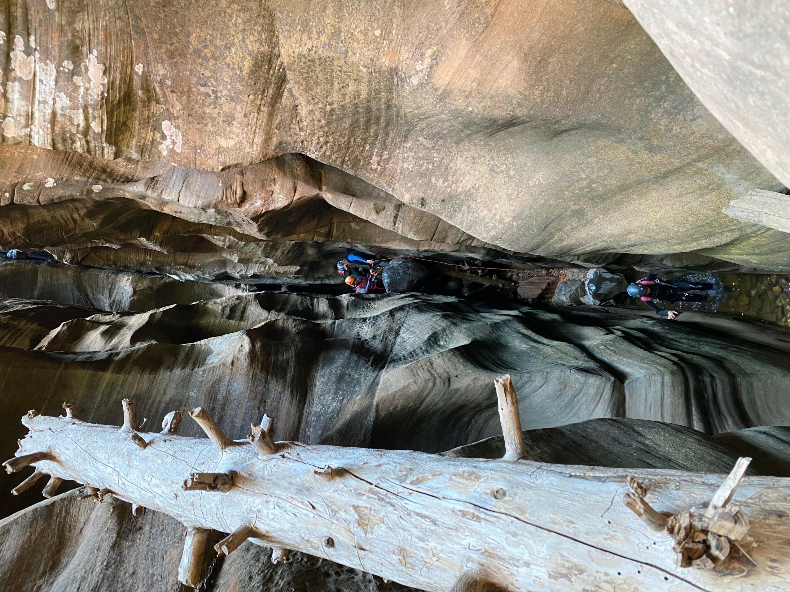 People exploring a narrow canyon with smooth, layered rock walls and a water-filled floor, some sitting on rocks or logs.