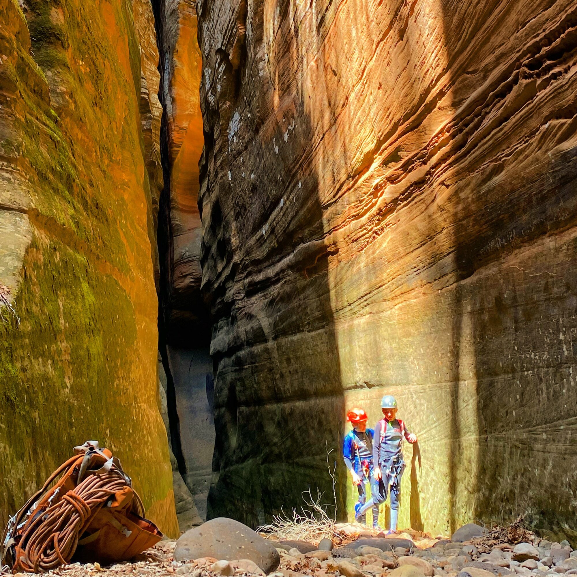 Two people wearing helmets and backpacks stand in a narrow canyon with tall, layered rock walls.