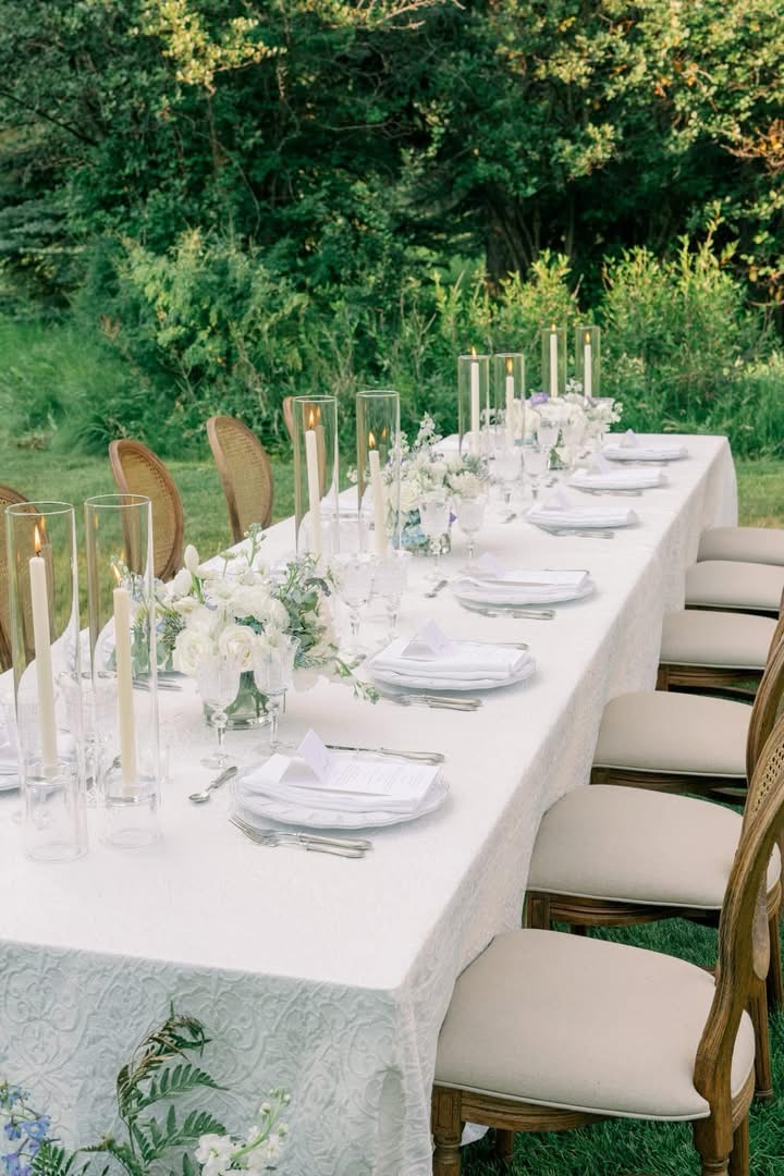 Long outdoor table set with white tablecloth, white plates, glasses, and floral centerpieces, surrounded by chairs, greenery in background.