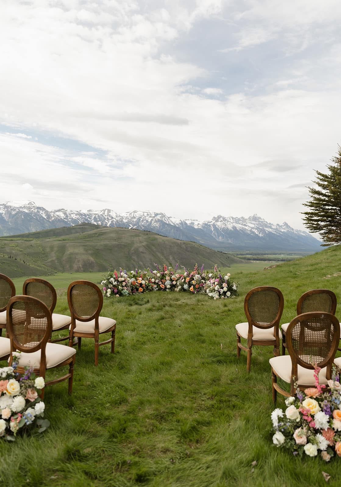 Outdoor wedding setup with chairs and floral arrangements on a grassy field, mountains and cloudy sky in background.