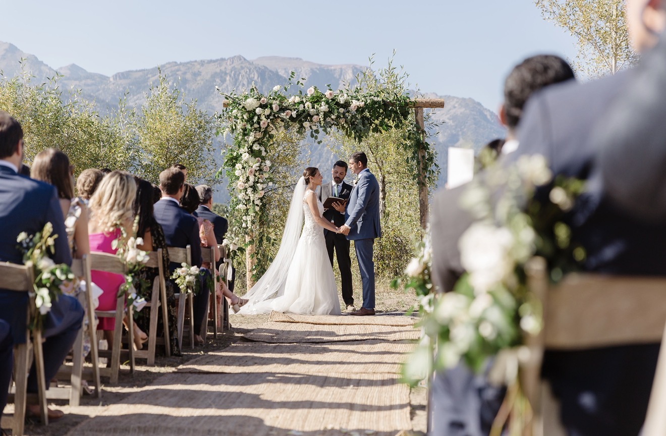Bride and groom holding hands during outdoor wedding ceremony with guests seated, mountains in background, sunny day.