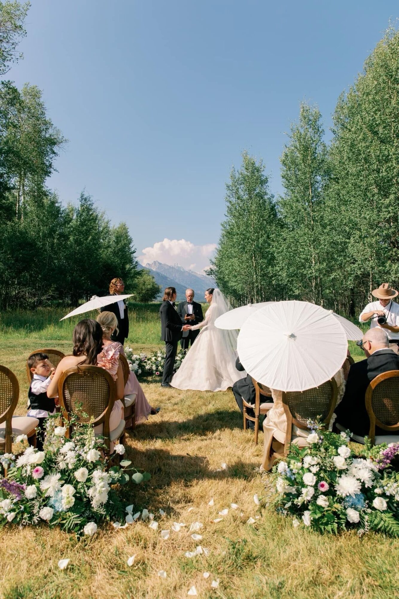 Outdoor wedding ceremony with guests seated on chairs, some holding umbrellas, surrounded by trees and flowers, under a clear blue sky.