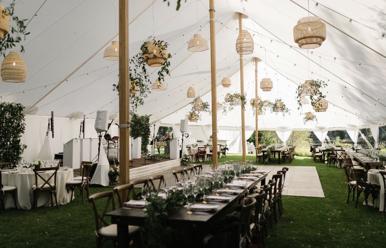 Indoor event space with tables, chairs, hanging lanterns, and floral decorations under a white tent ceiling.