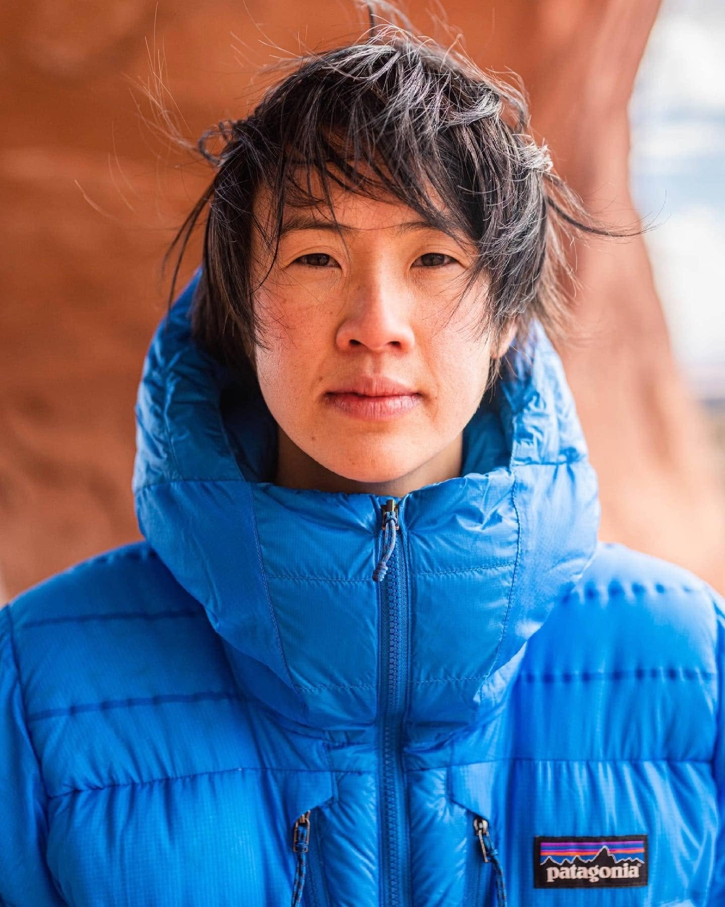 Person with short black hair wearing a bright blue puffer jacket outdoors, background of reddish-brown rock formations.