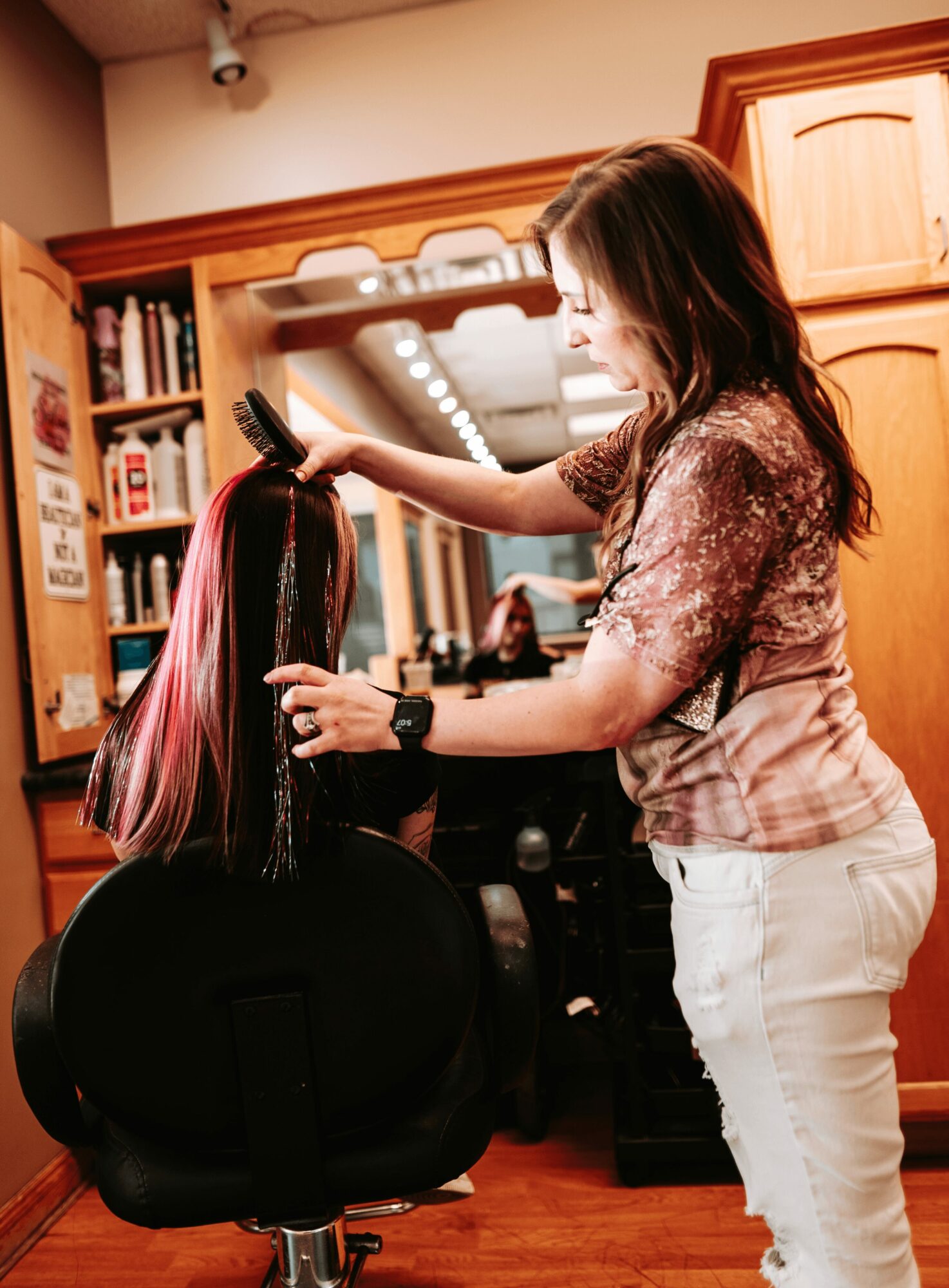 Woman with long hair getting her hair styled in a salon, facing mirror, with hairdresser working on her hair.