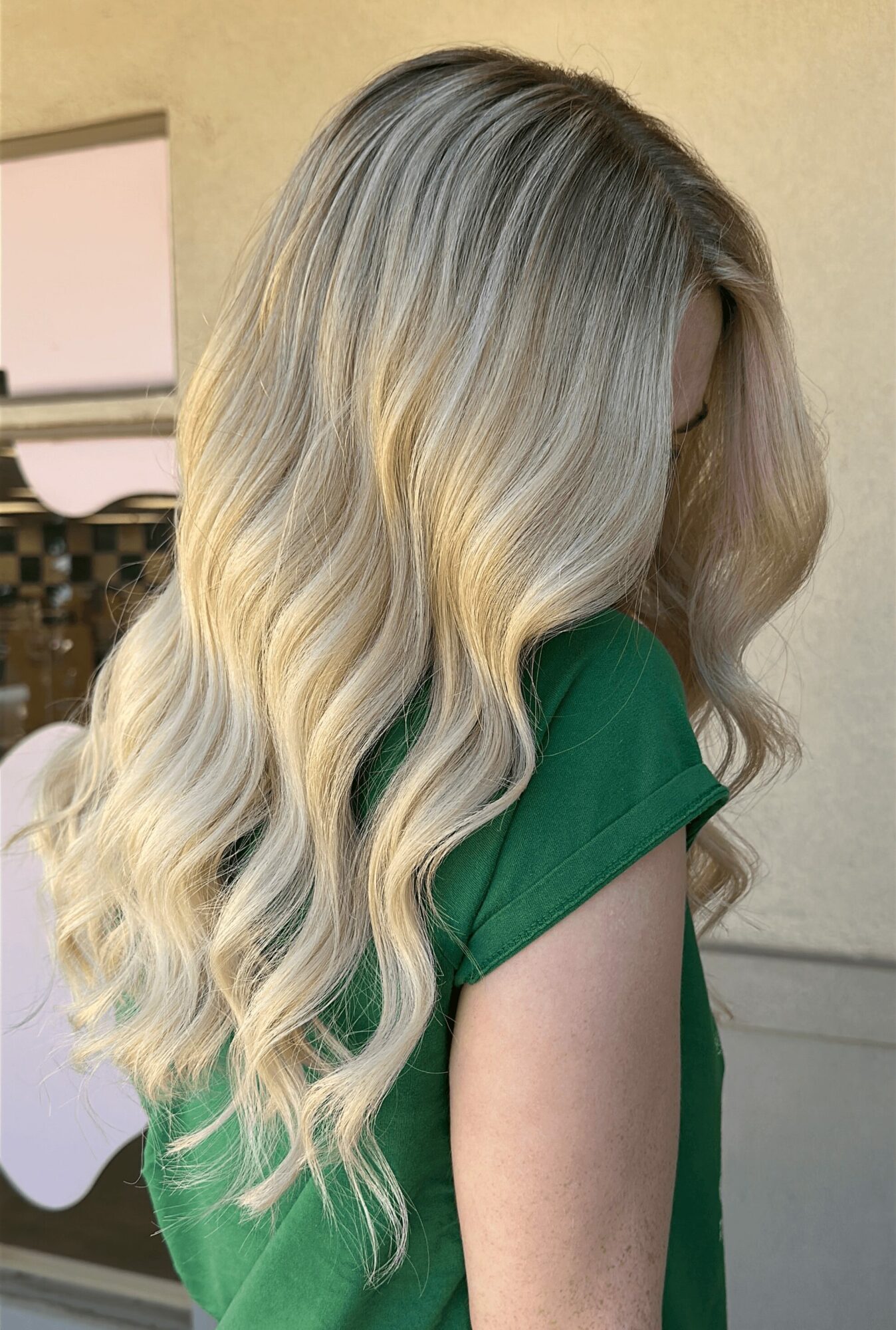 Side view of a woman with long, wavy blonde hair wearing a green top, standing indoors.