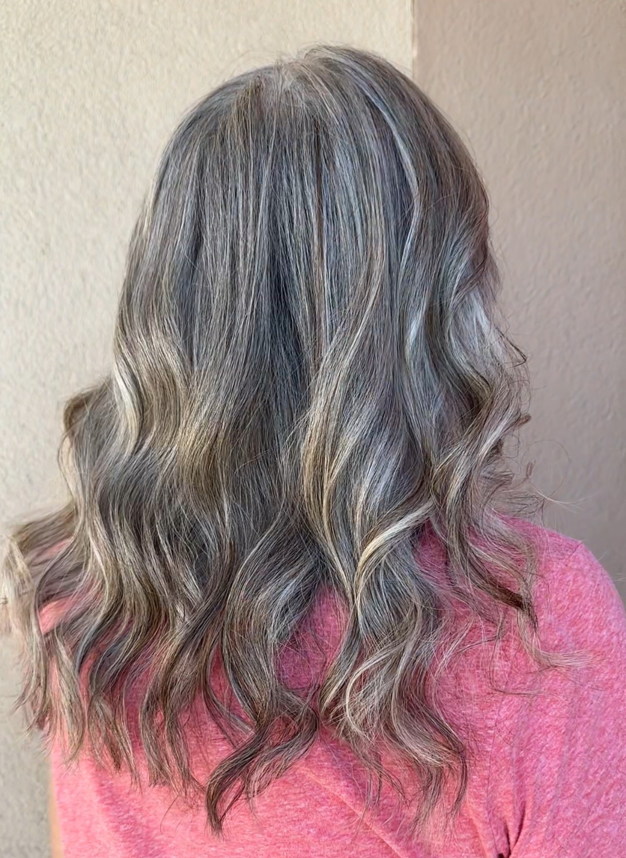 Back view of wavy, shoulder-length hair with blonde and gray highlights, wearing a pink top.