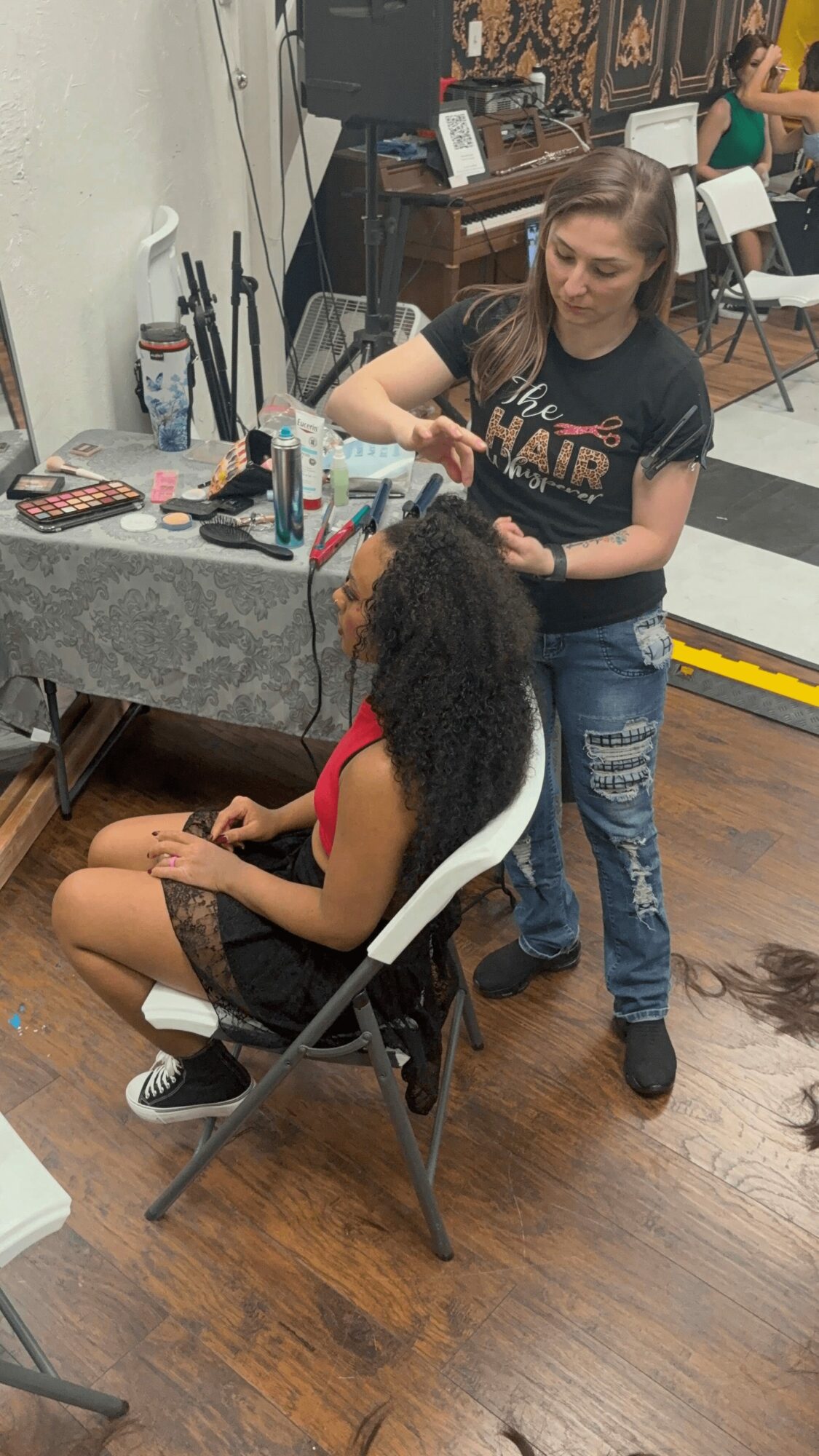 A woman with long hair and a black T-shirt styles a seated person with curly hair in a salon, with styling tools on a table nearby.