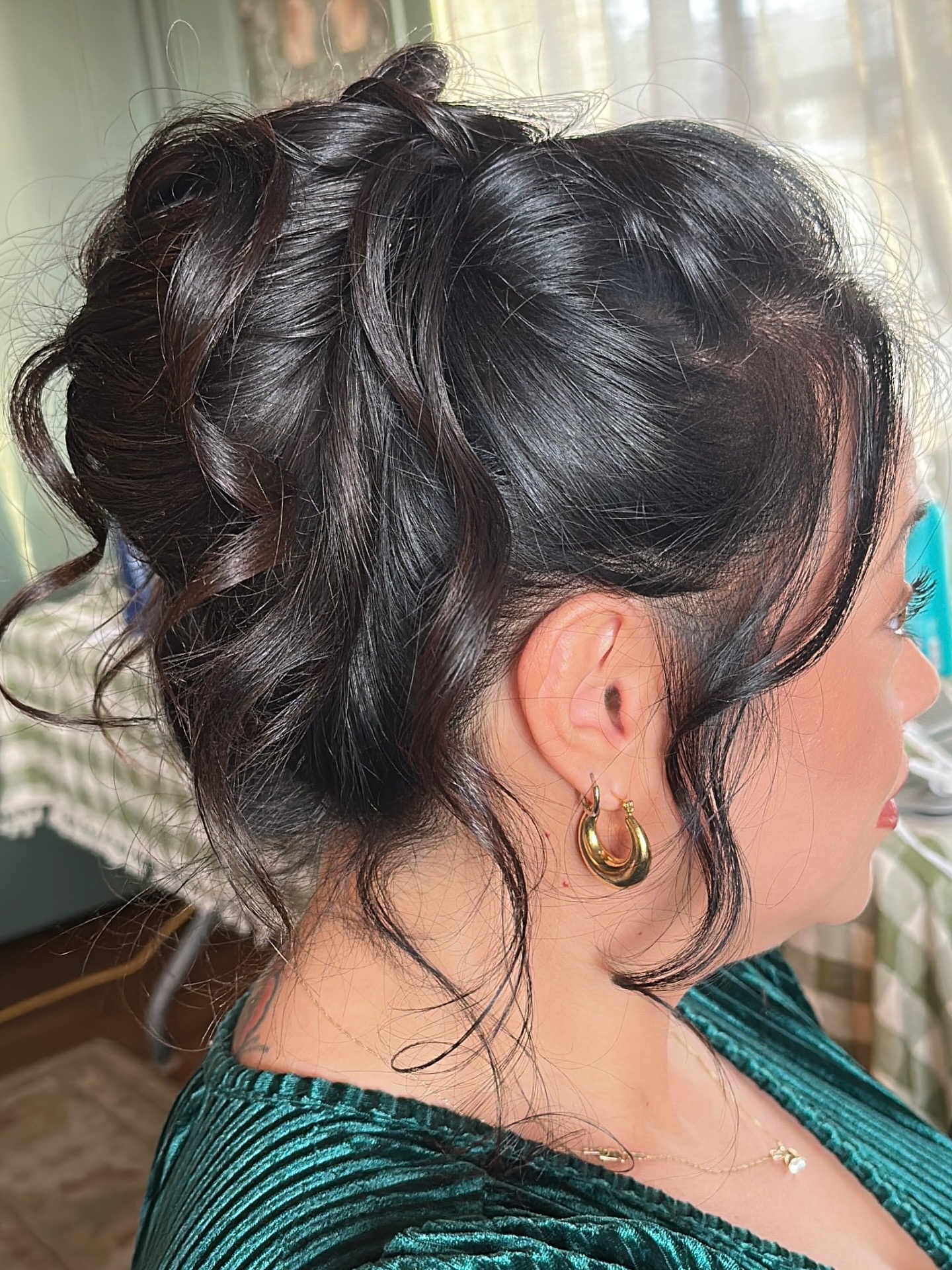 Side view of a woman with dark, curly hair styled in an updo, wearing gold hoop earrings and a green top.