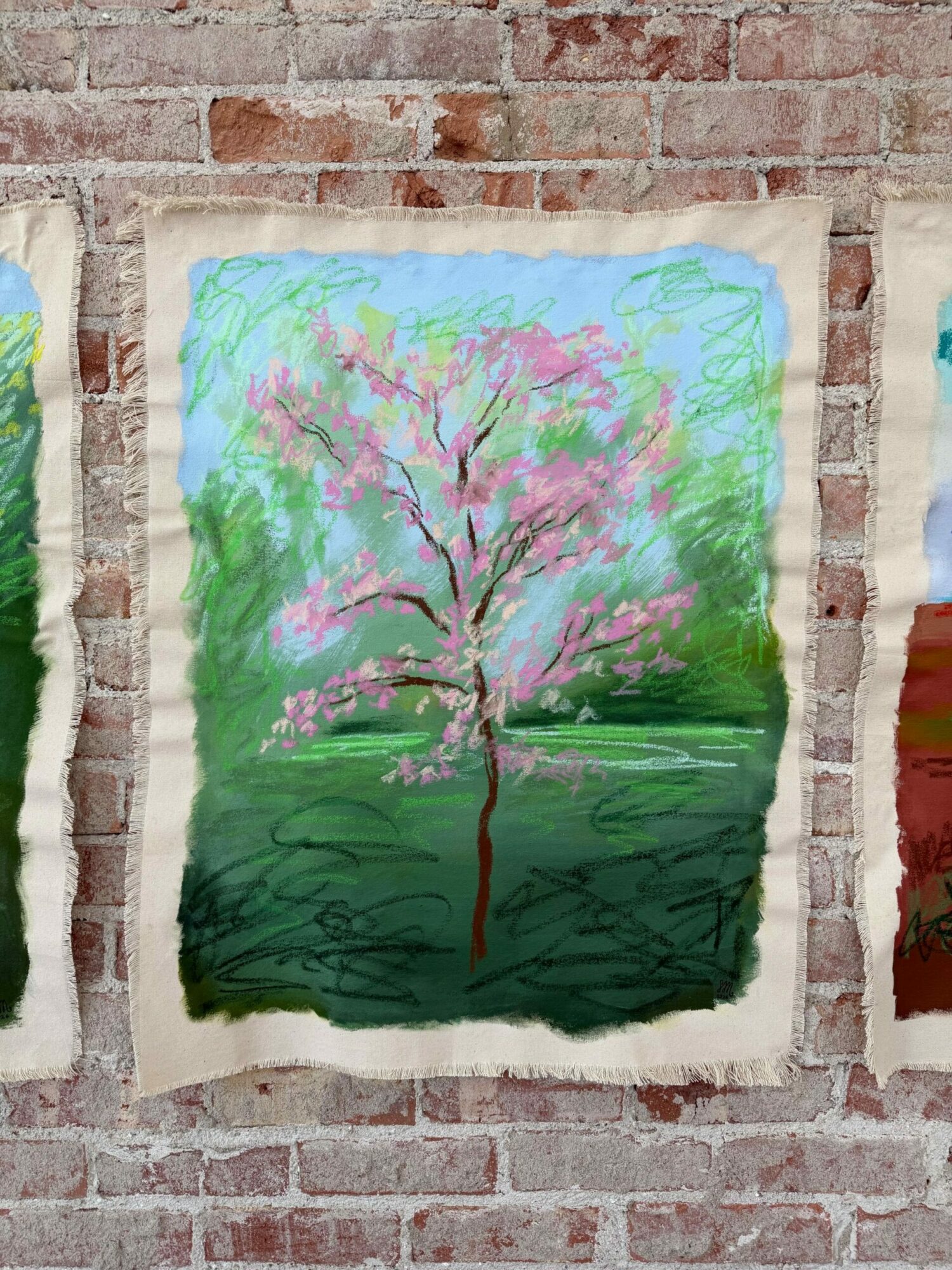 Painting of a blooming tree with pink flowers, green grass, and a blue sky, hanging on a brick wall.