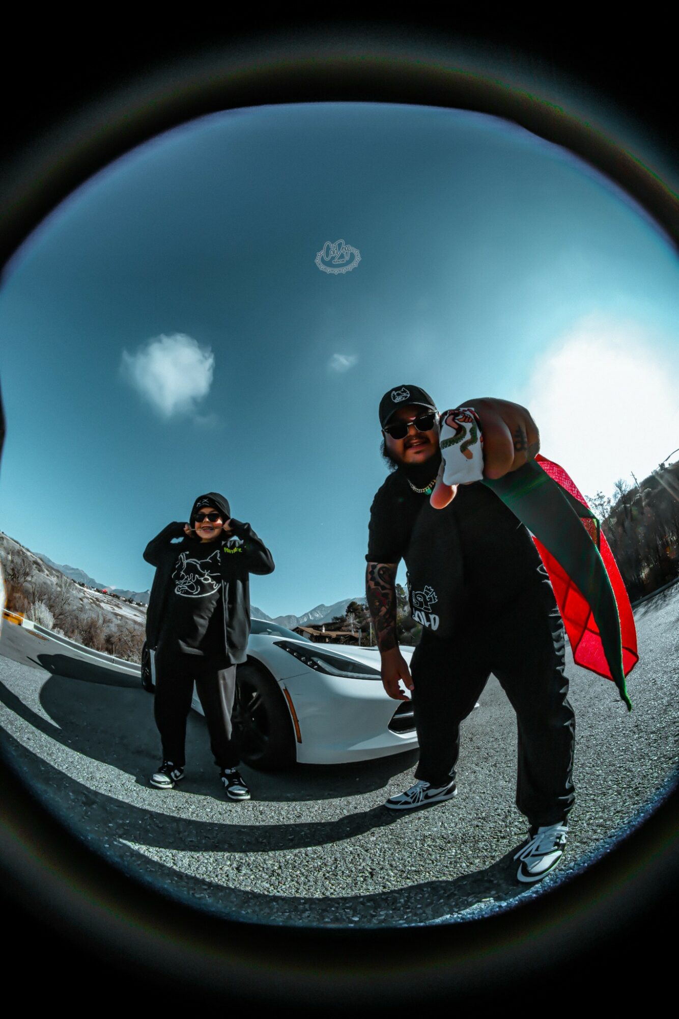 Three people standing outdoors near a car under a blue sky with clouds, one holding a backpack, fisheye lens effect.