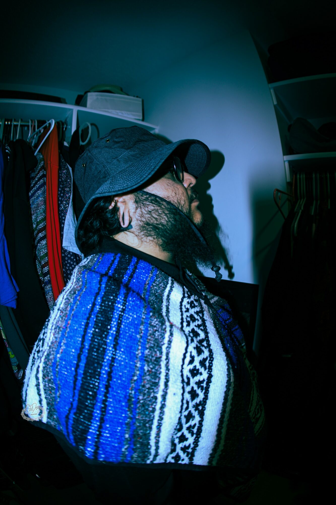 Person with a beard, glasses, and a hat, wearing a patterned sweater, sitting in a room with clothes and furniture.