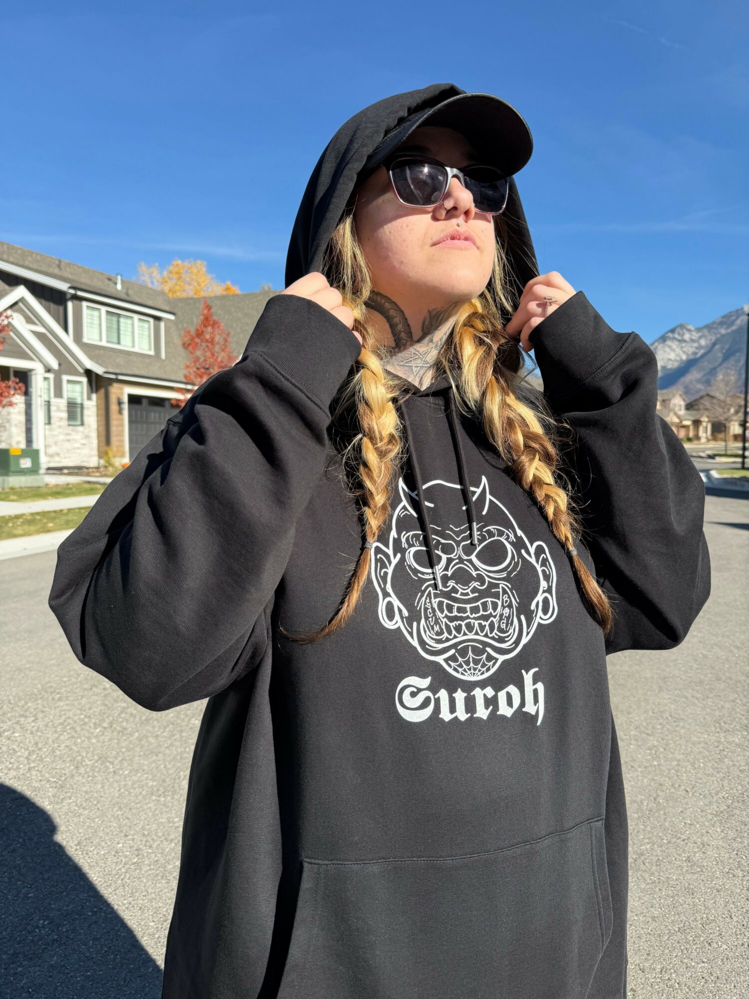Young woman with braided hair wearing sunglasses and a hoodie, standing outdoors on a sunny day.