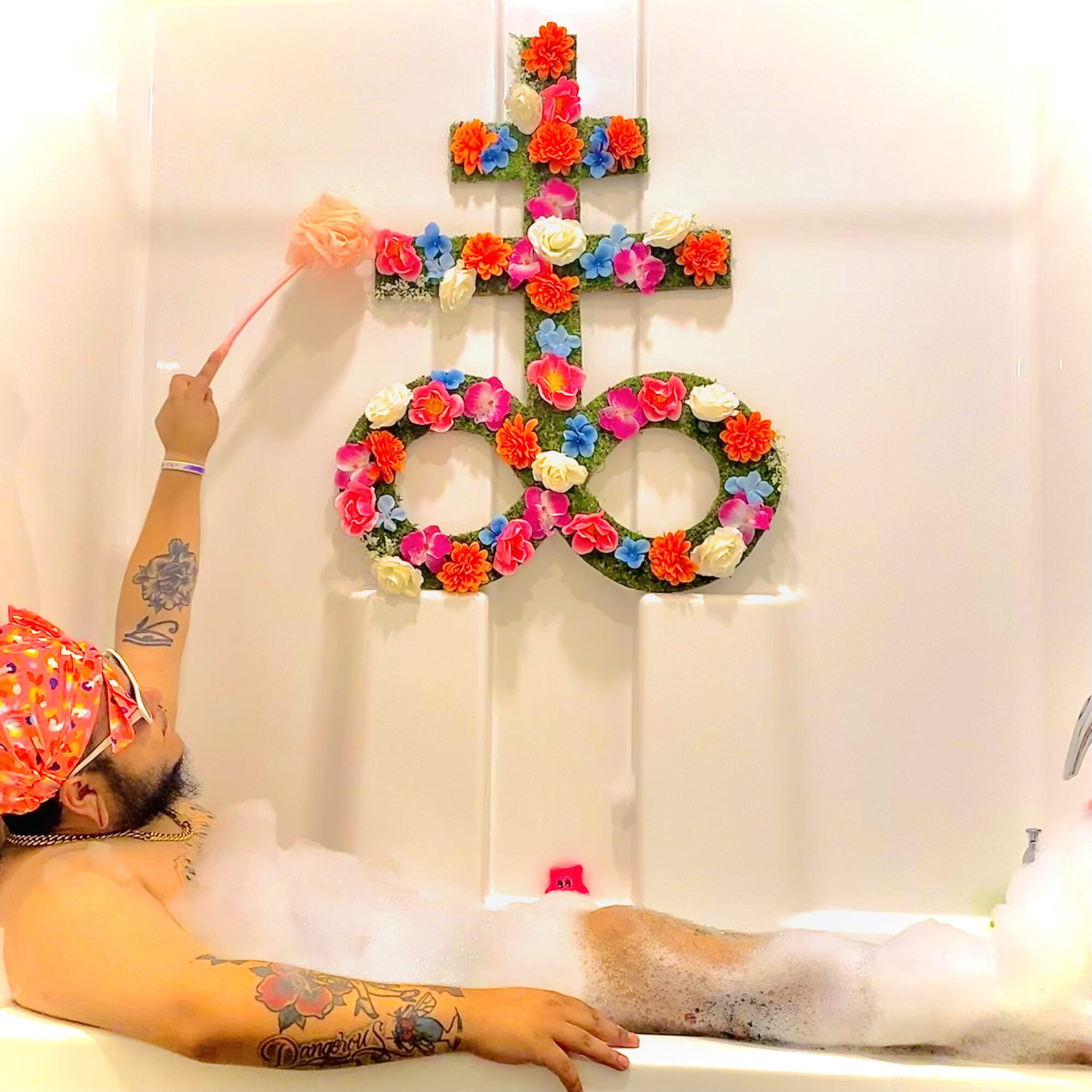 Person with tattoos in a bathtub, cleaning a floral cross and number decoration on the wall, with bubbles and foam present.