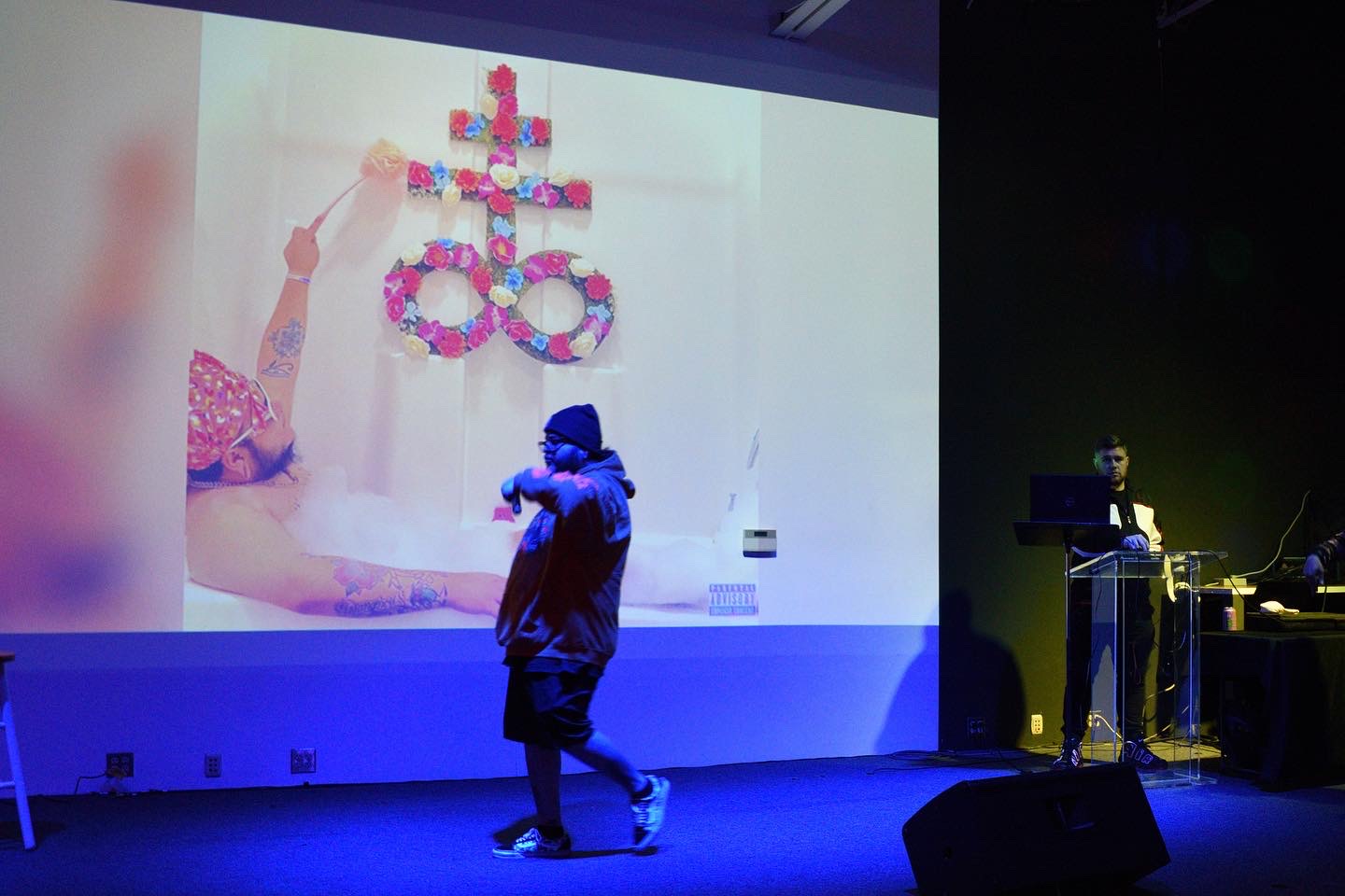 Person standing in front of a large screen displaying a colorful craft project with flowers and a figure's hand, in a dark room.