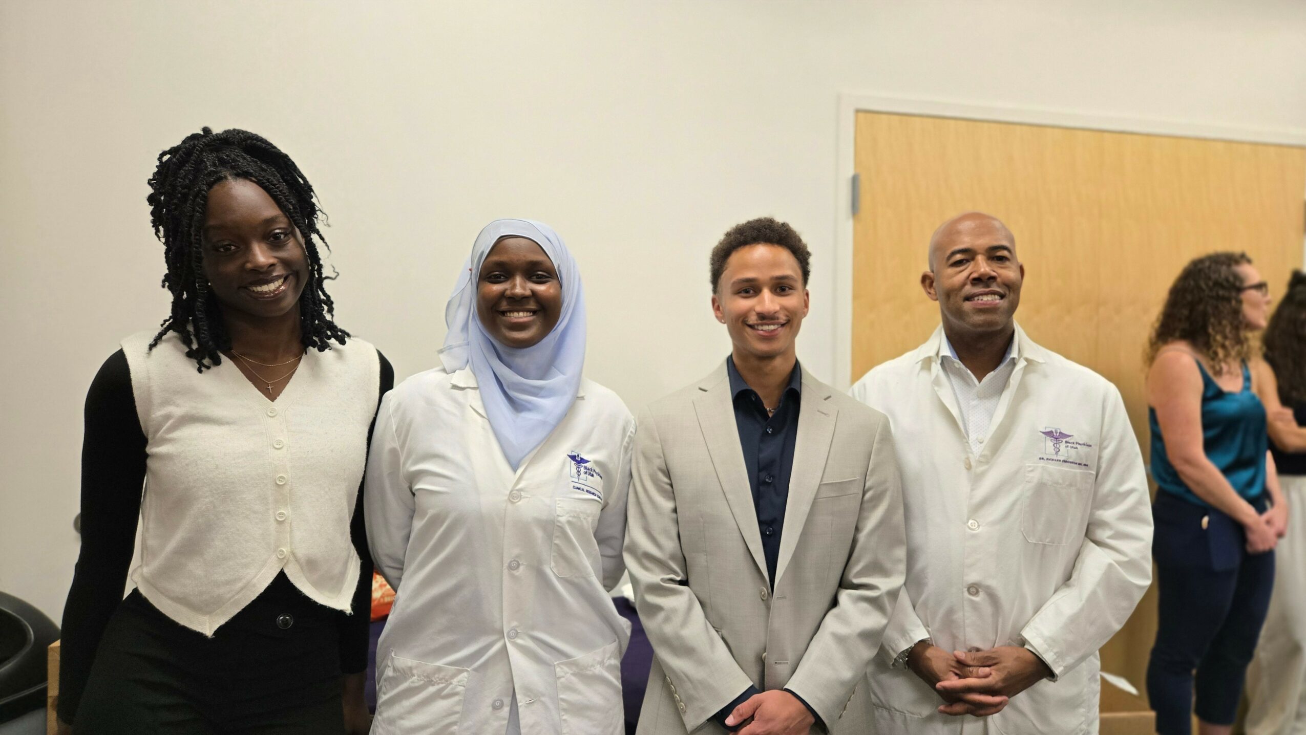 Four people standing together, smiling, in a room with a plain wall and a door in the background.