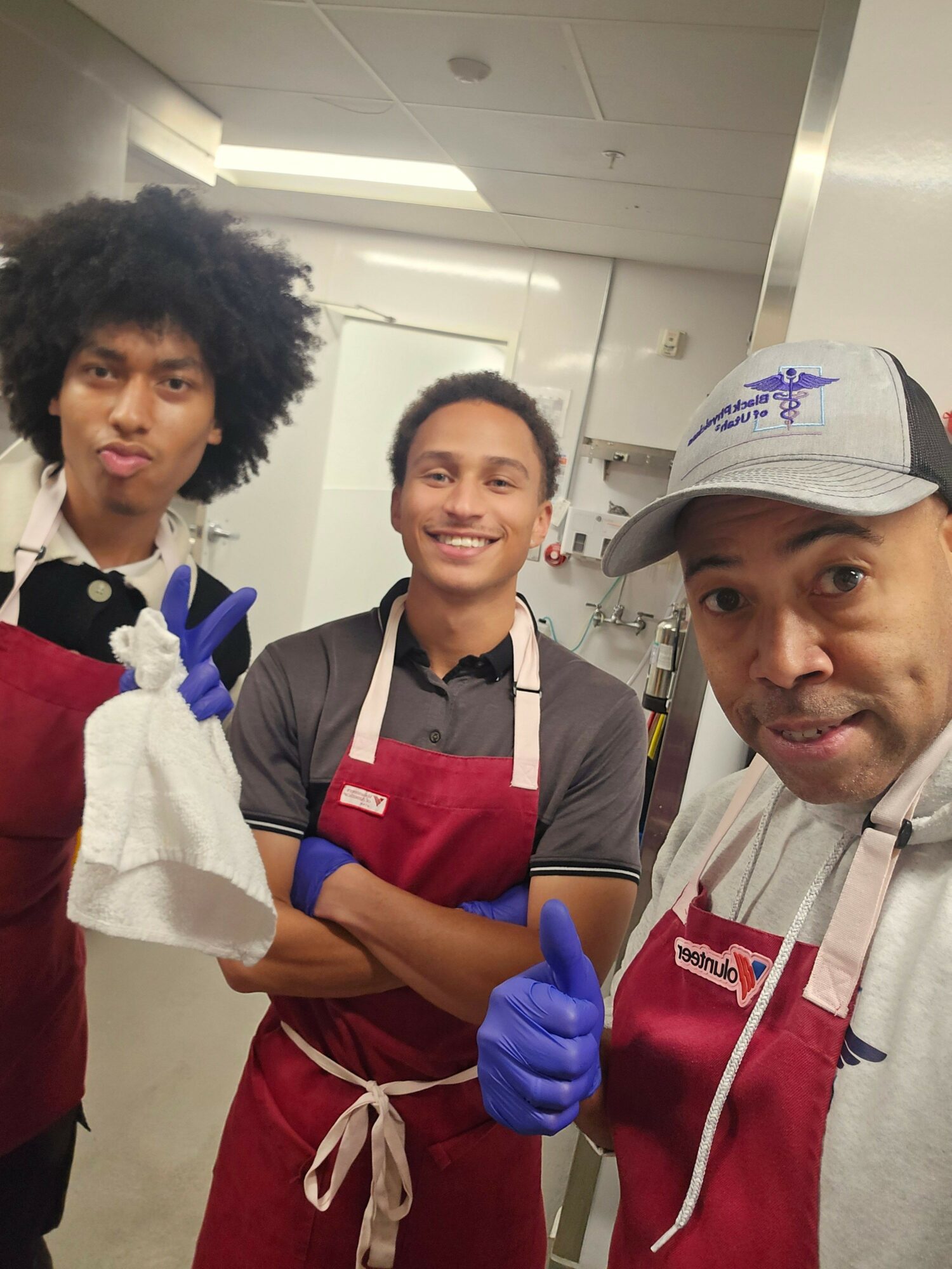 Three people in a kitchen, two wearing aprons and gloves, one in a cap, smiling at the camera.
