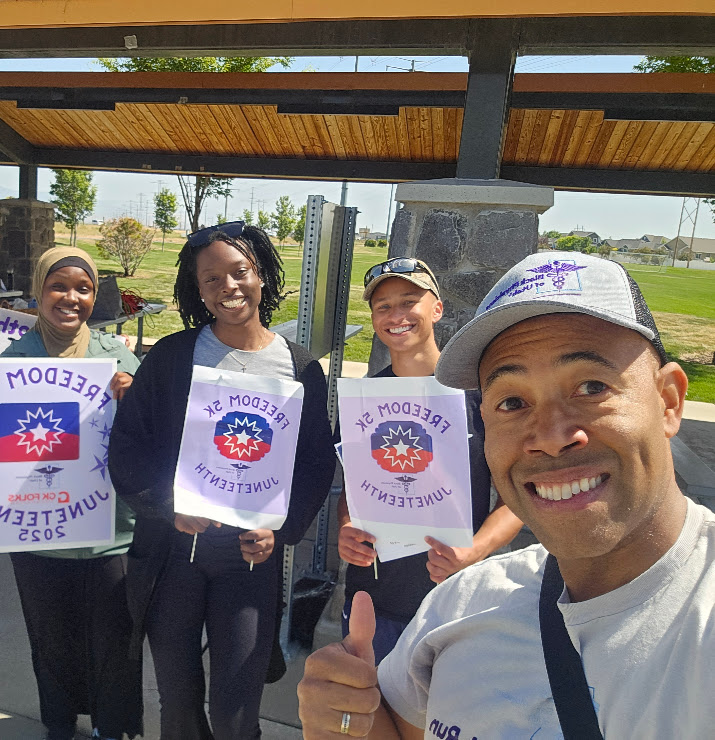 Four people outdoors holding signs with a logo and text, smiling, under a wooden structure with a park in the background.