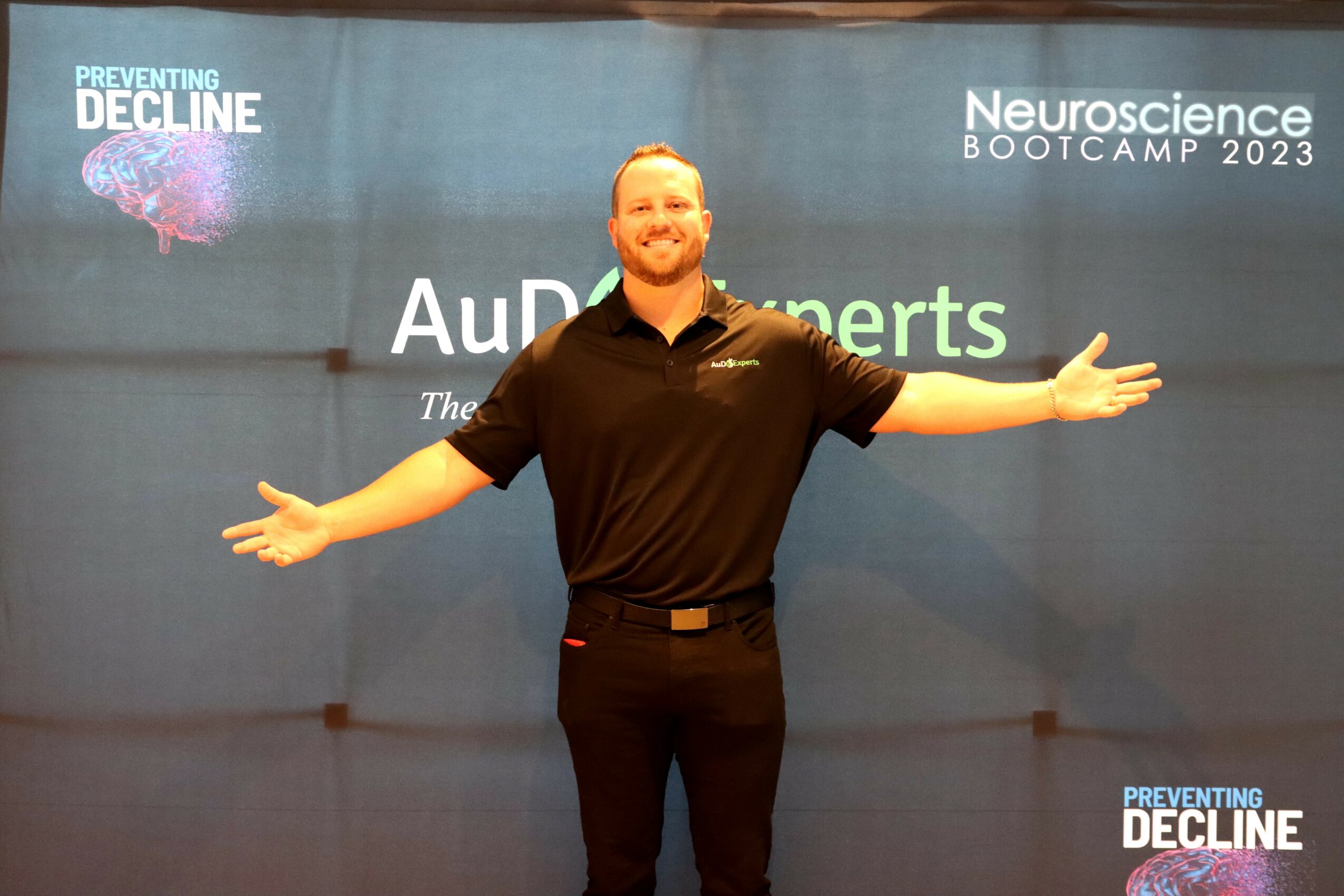 Man with arms outstretched standing in front of a blue backdrop with event text, smiling.