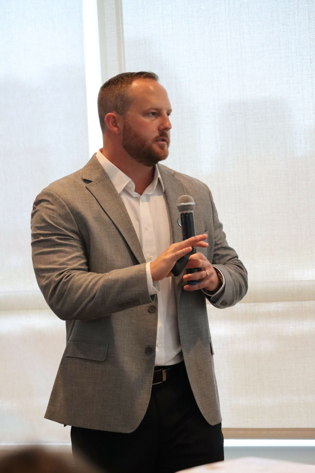Man in a gray blazer and white shirt holding a phone, standing indoors near a window.