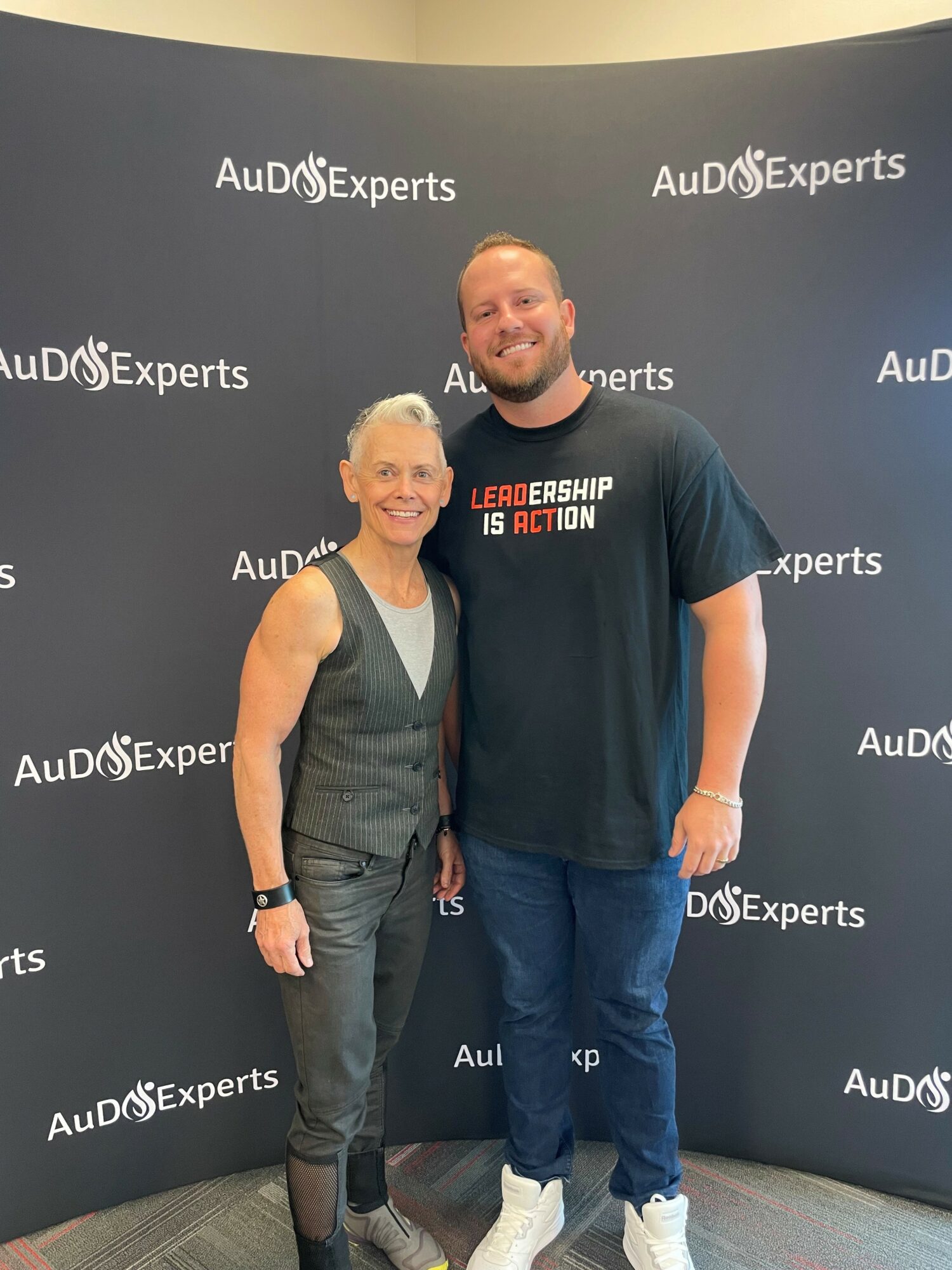 Two people standing together in front of a black backdrop with 'AuD@Experts' logo, smiling, one in a gray vest and the other in a black T-shirt.