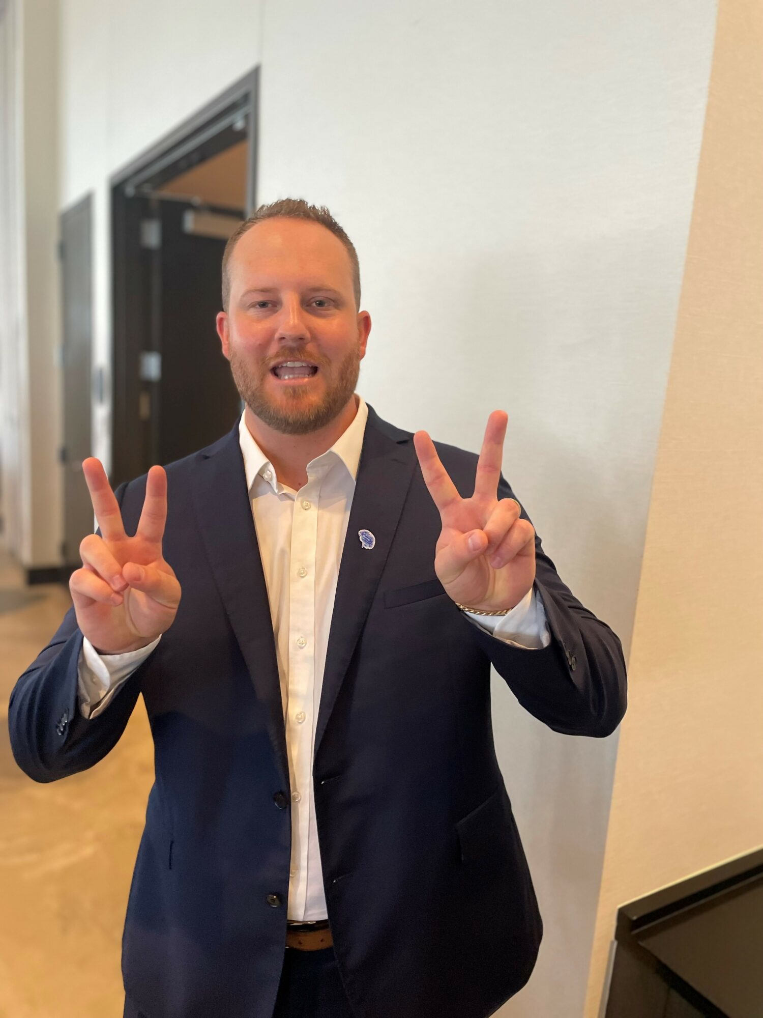 Man in suit making peace signs with both hands, smiling, standing indoors against a white wall.