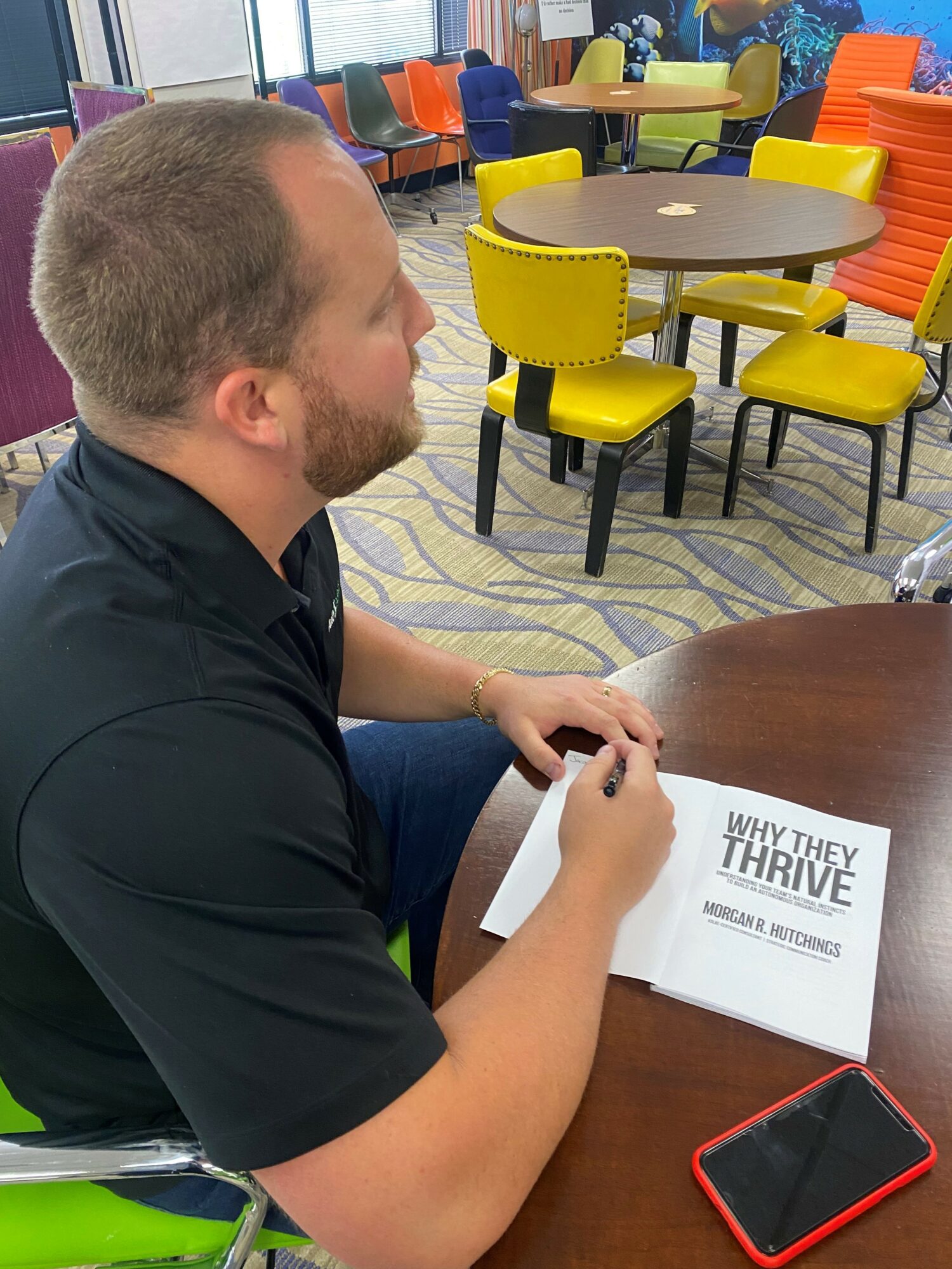 Man sitting at a table in a colorful room, holding a paper titled 'WHY THEY THRIVE' with a red phone nearby.