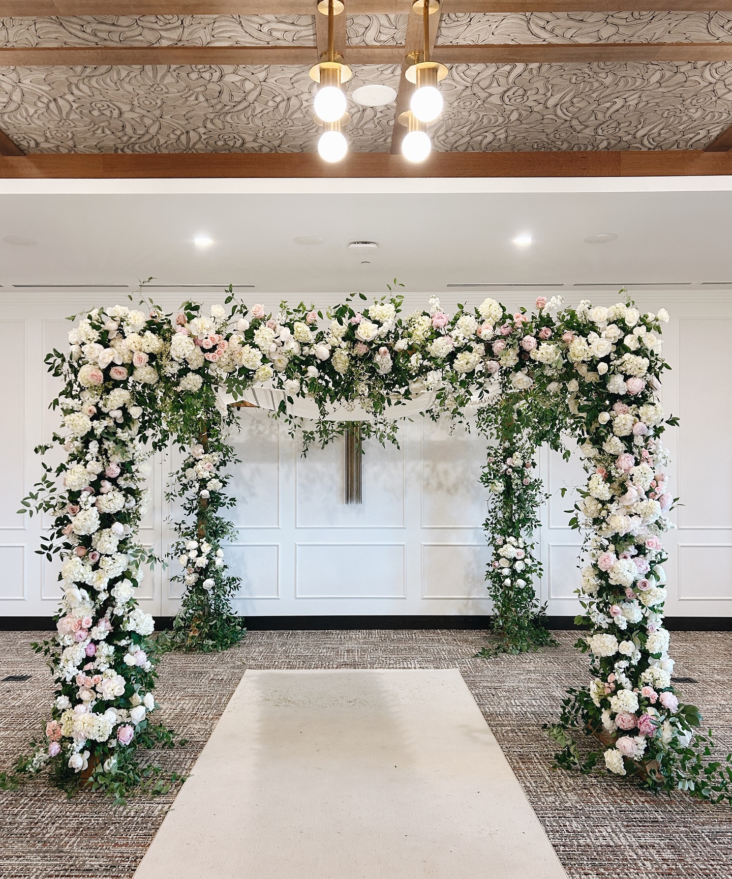 Floral arch with white and pink flowers and greenery, set on a carpeted floor with a white aisle in front.