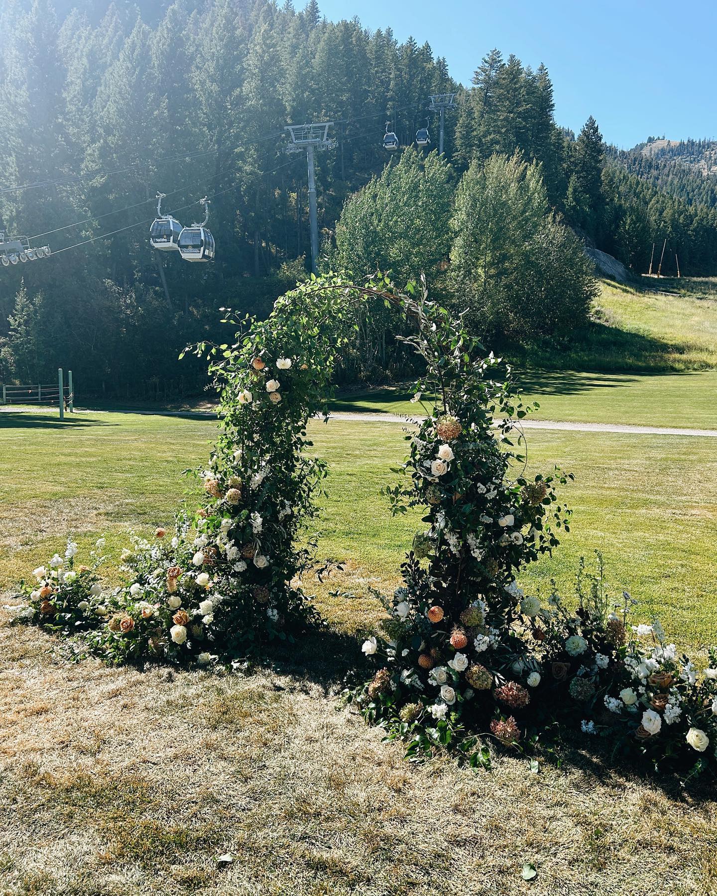 Flower arch with white and pink roses on grassy area, trees and ski lift in background, clear blue sky.