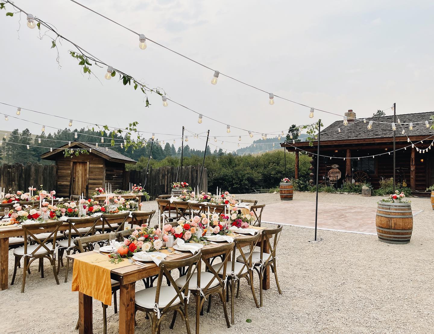 Outdoor event setup with decorated tables, string lights, and barrels, in a rustic setting with a building and trees in background.