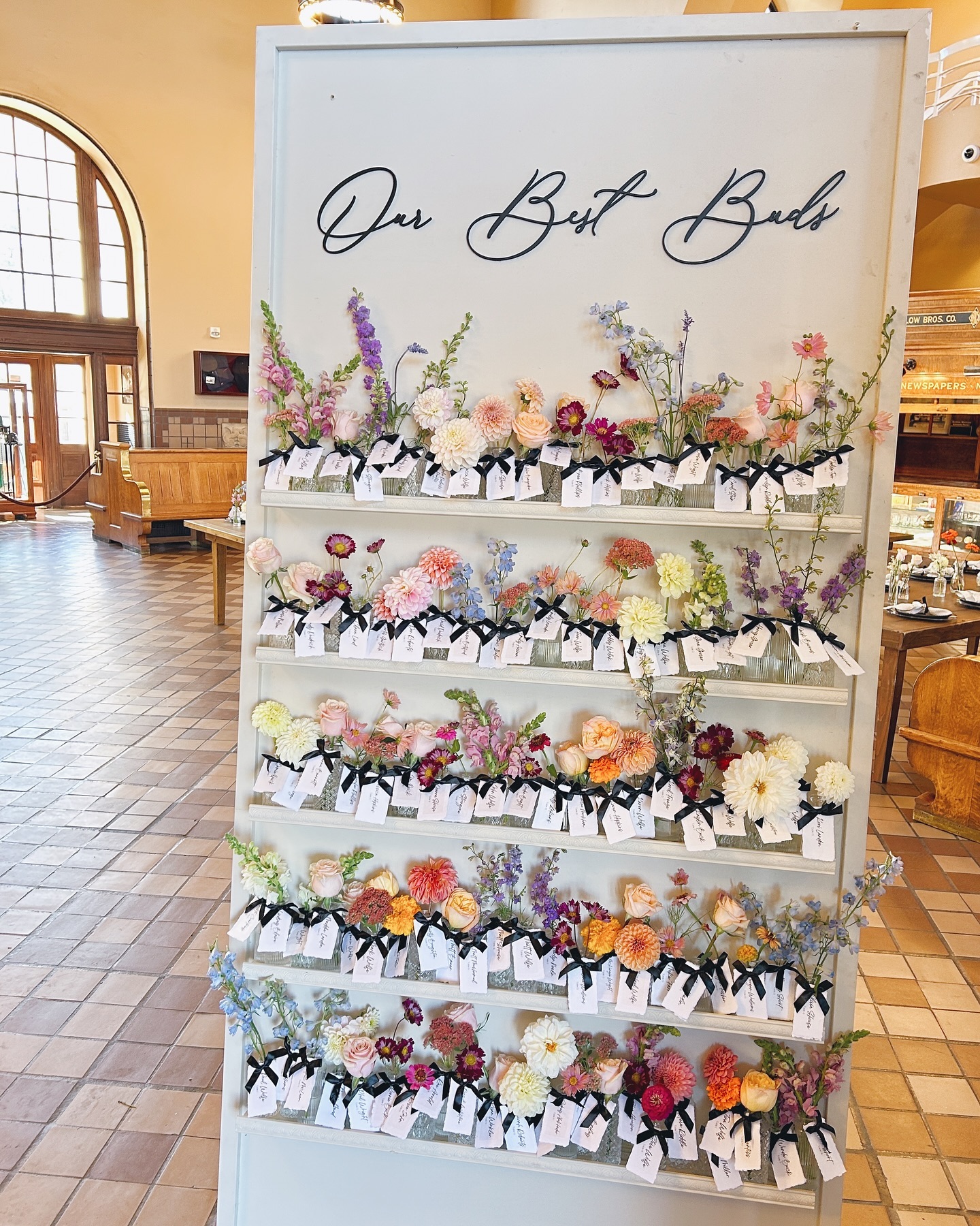 Whiteboard with handwritten text and colorful flowers arranged on shelves, in a spacious room with large windows and wooden furniture.