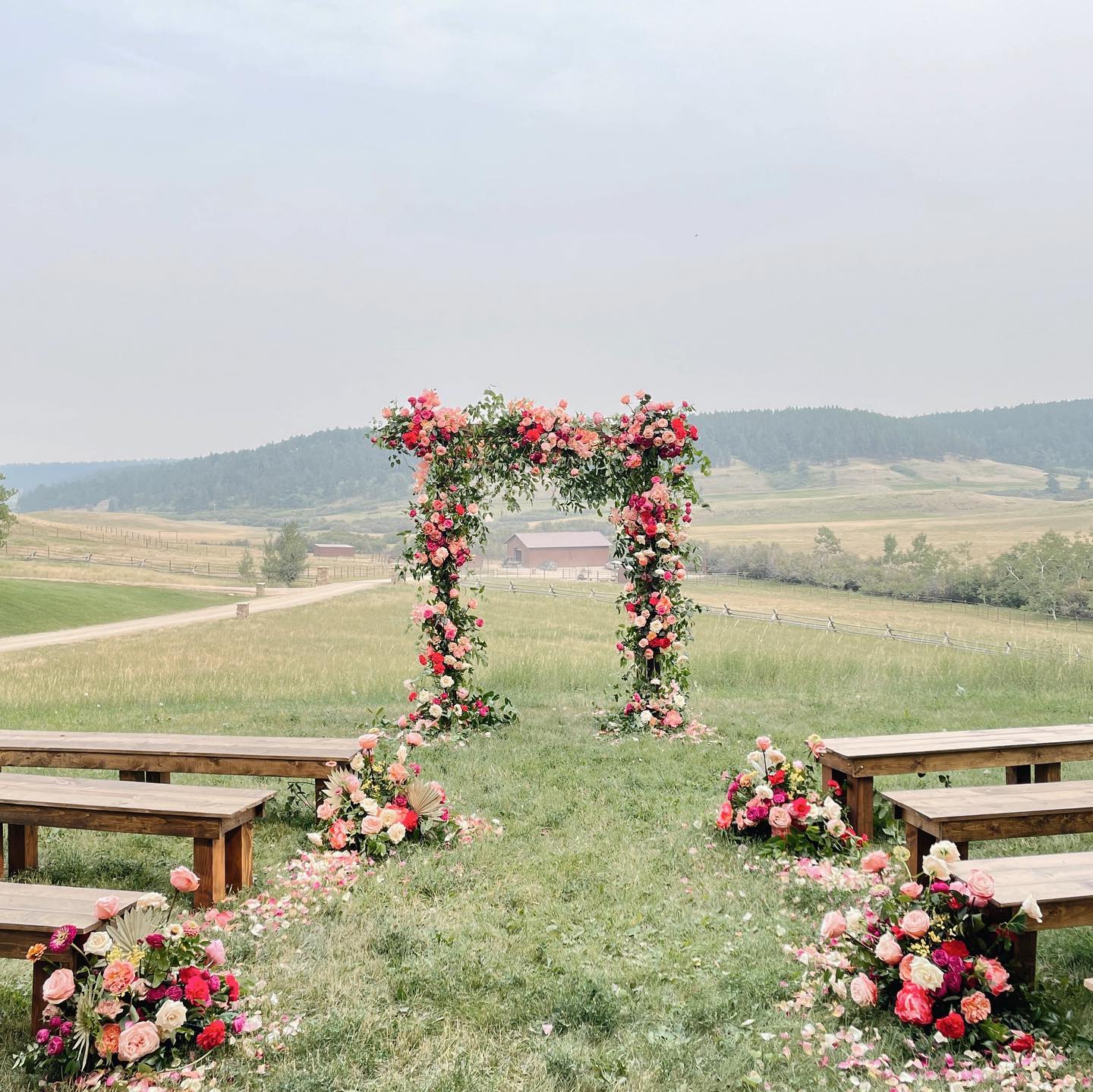 Outdoor wedding setup with floral arch, benches, and flower arrangements on grass, overlooking rolling hills and cloudy sky.