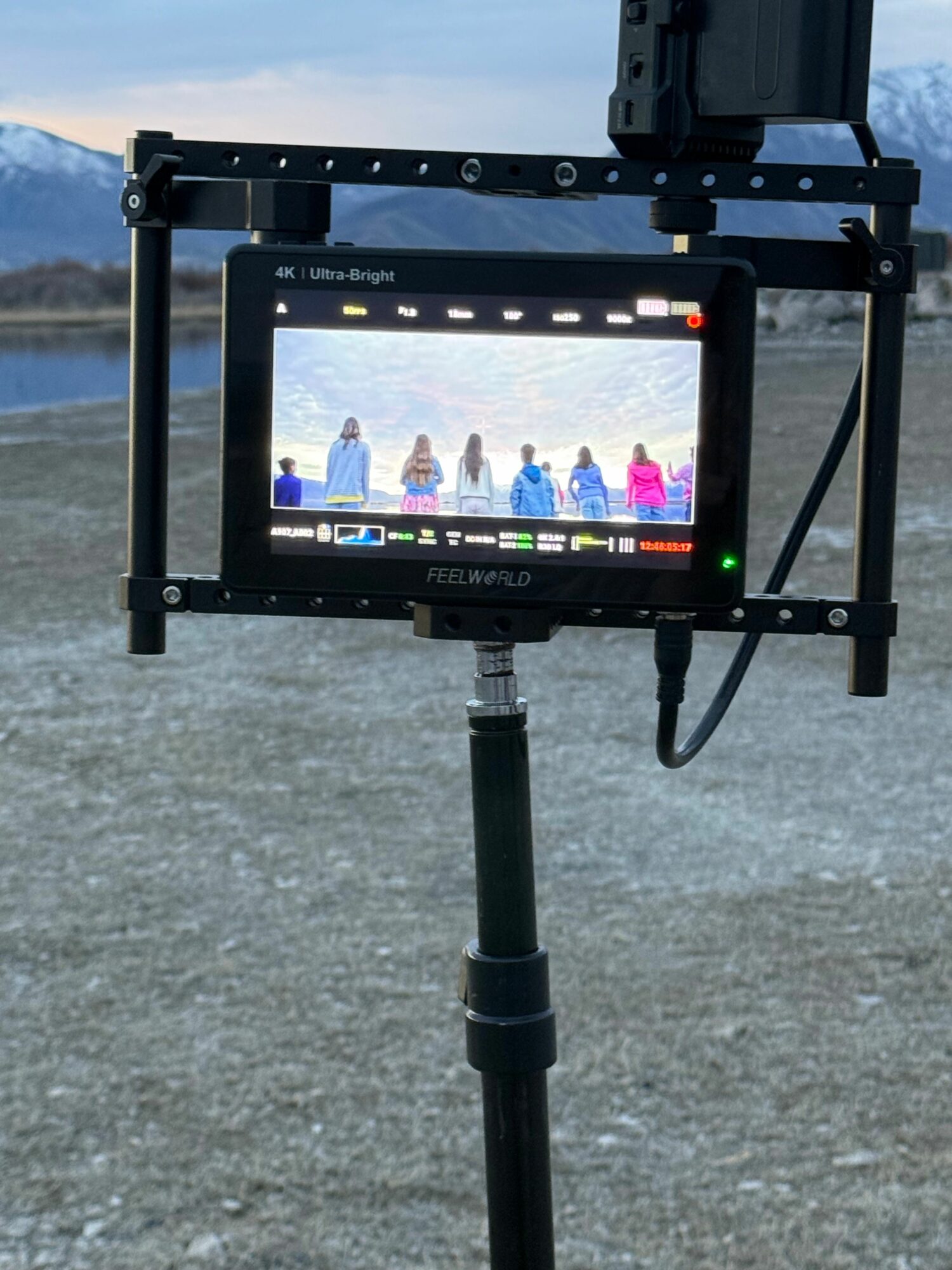 A camera monitor mounted on a rig, displaying a scene with multiple people outdoors, with a textured ground surface.