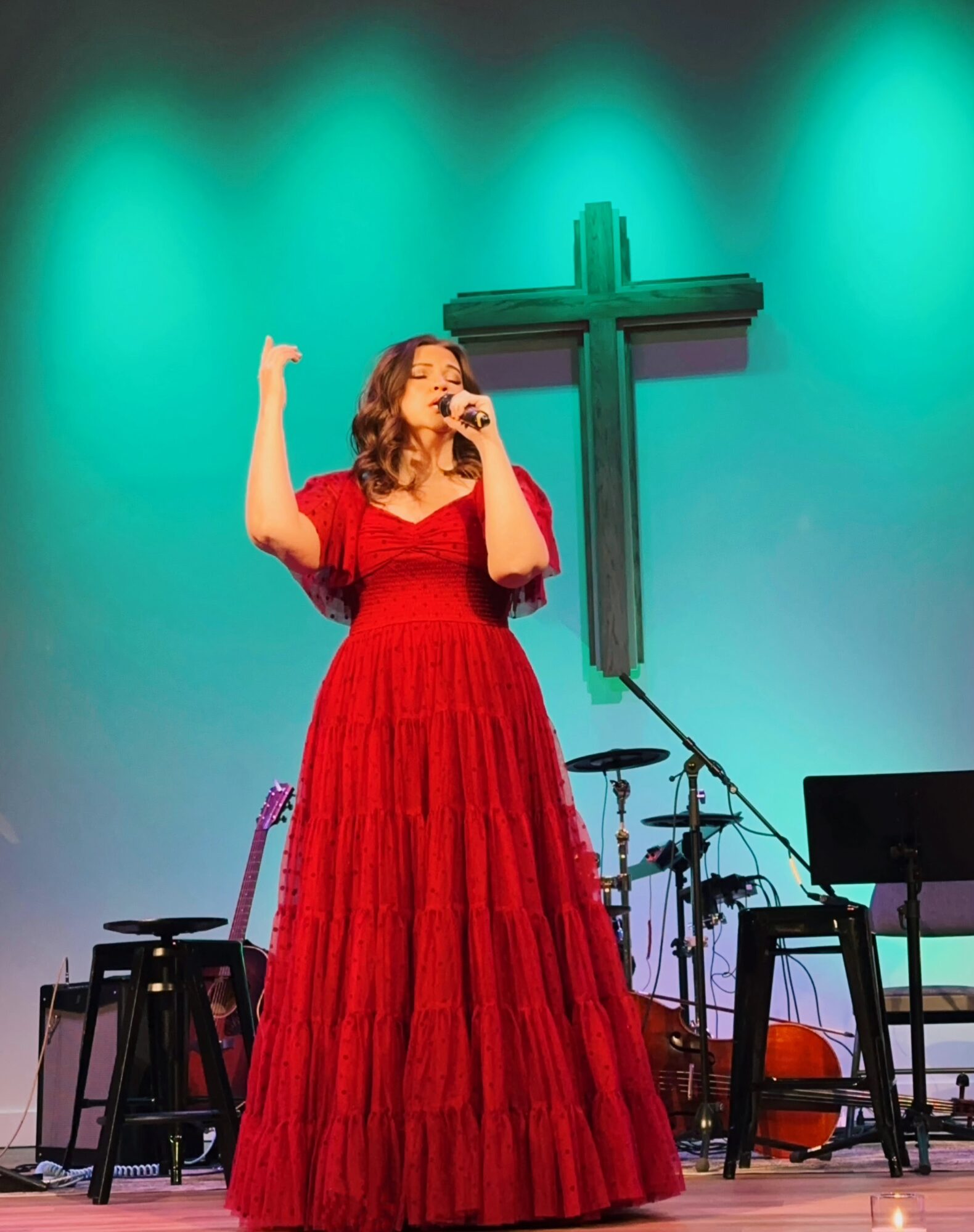 Woman in red dress singing on stage with a cross in the background, musical instruments around her.
