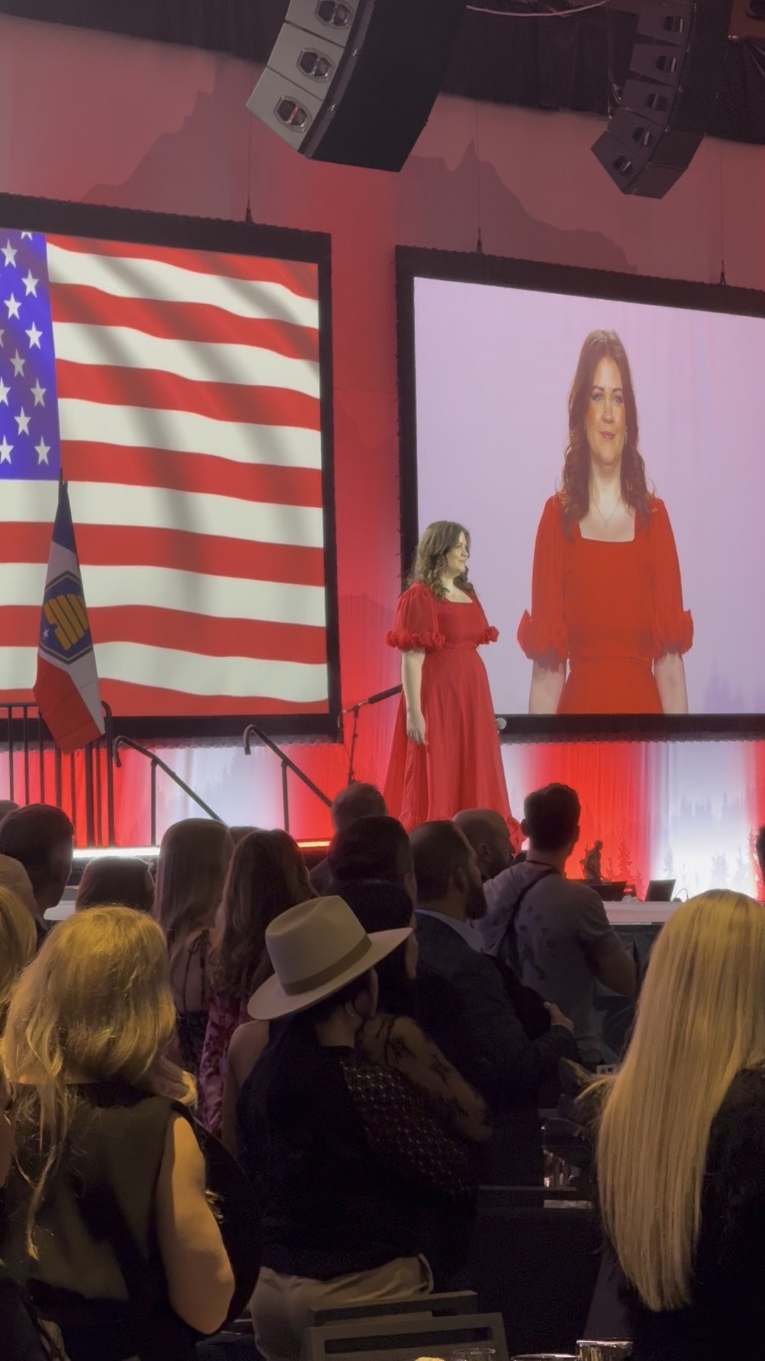 Woman in red dress speaking on stage with large American flag and screen showing her image, audience seated, some wearing hats.