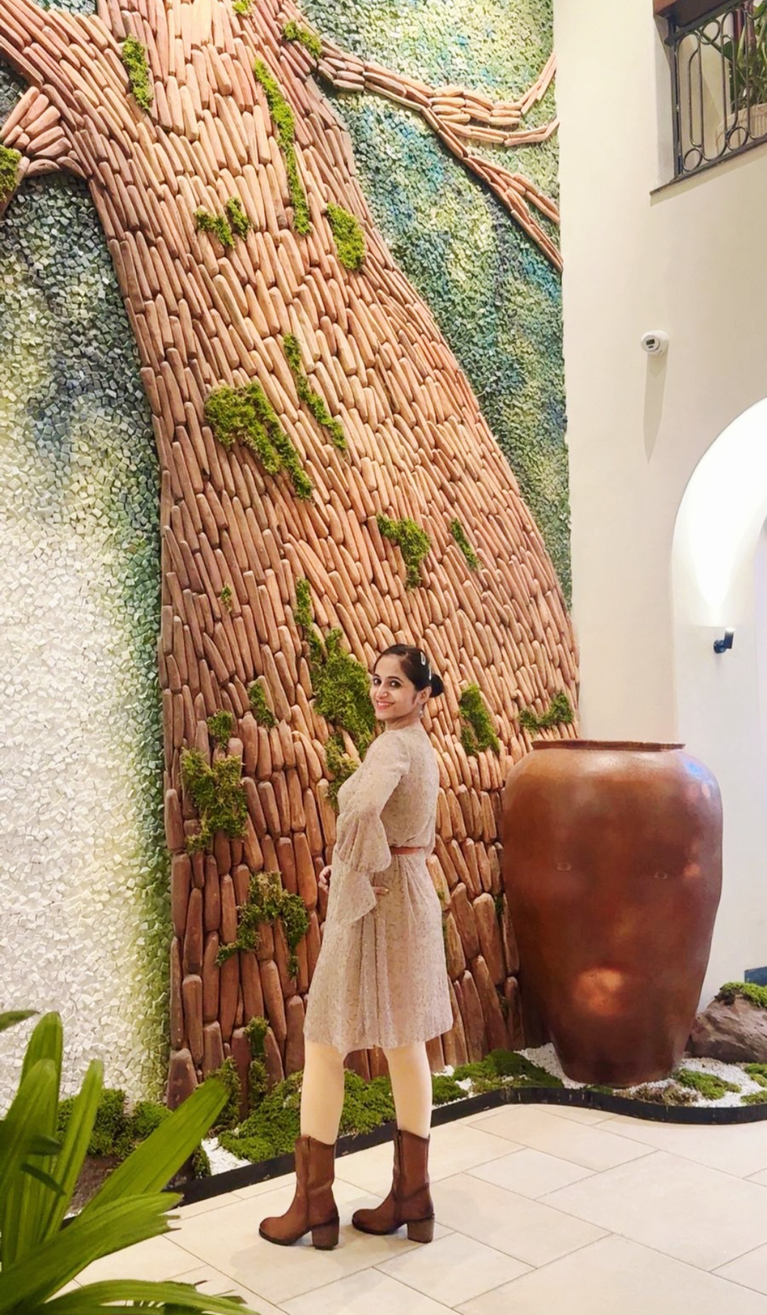 Woman in beige dress and boots standing indoors near a large clay pot and a textured wall with a vertical garden.