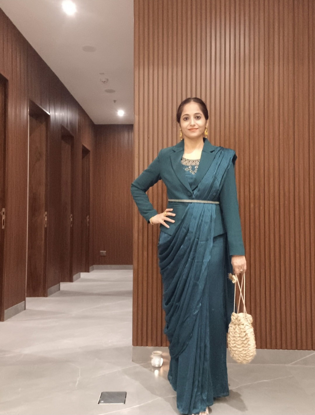 Woman in teal saree standing in a hallway with wooden wall panels, smiling, holding a beige bag, with a neutral background.