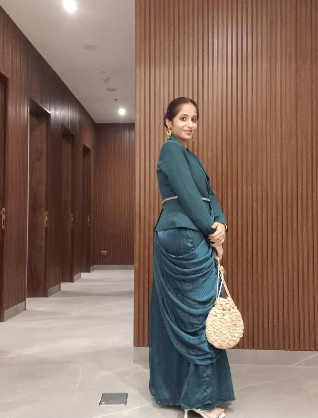 Woman in teal dress holding a woven bag, standing against a wooden-paneled wall in an indoor corridor.