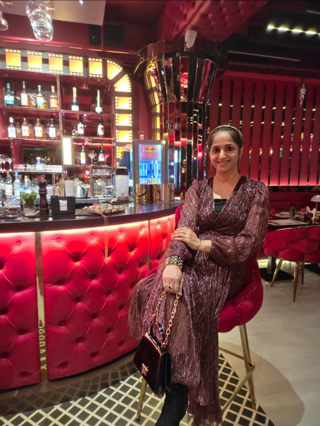 Woman in a patterned dress sitting on a red barstool in a brightly lit bar with red and gold decor.